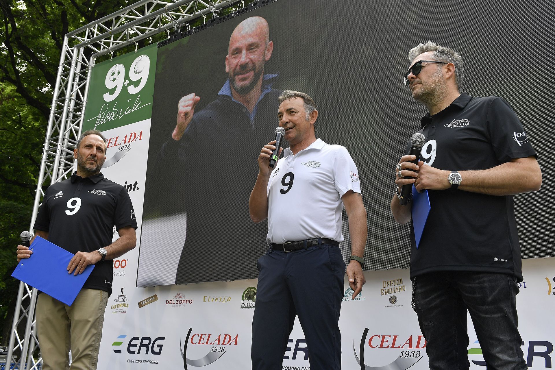 Tre uomini sul palco; uno parla al microfono. Un grande schermo mostra un uomo. Evento all'aperto con i loghi degli sponsor.