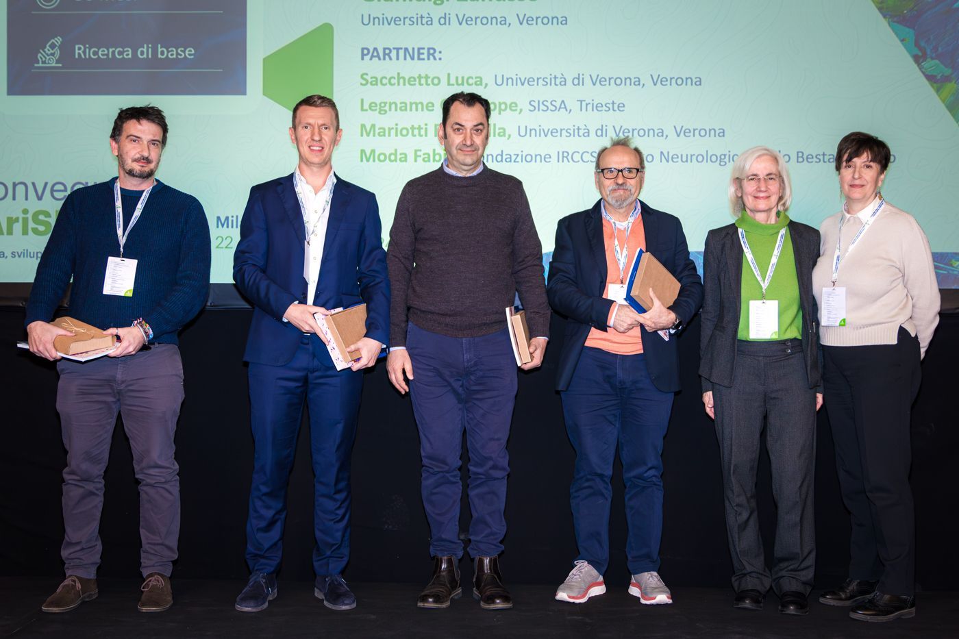 Sei persone in piedi su un palco, con in mano dei libri, davanti allo striscione di una conferenza.