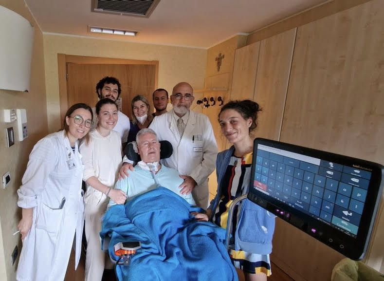 Gruppo di persone in una stanza d'ospedale con un paziente a letto, circondato dal personale medico.