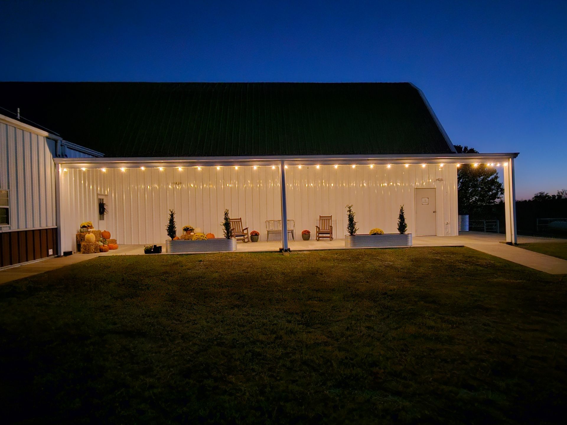 A barn with string lights, pumpkins, and seating on a patio at dusk.