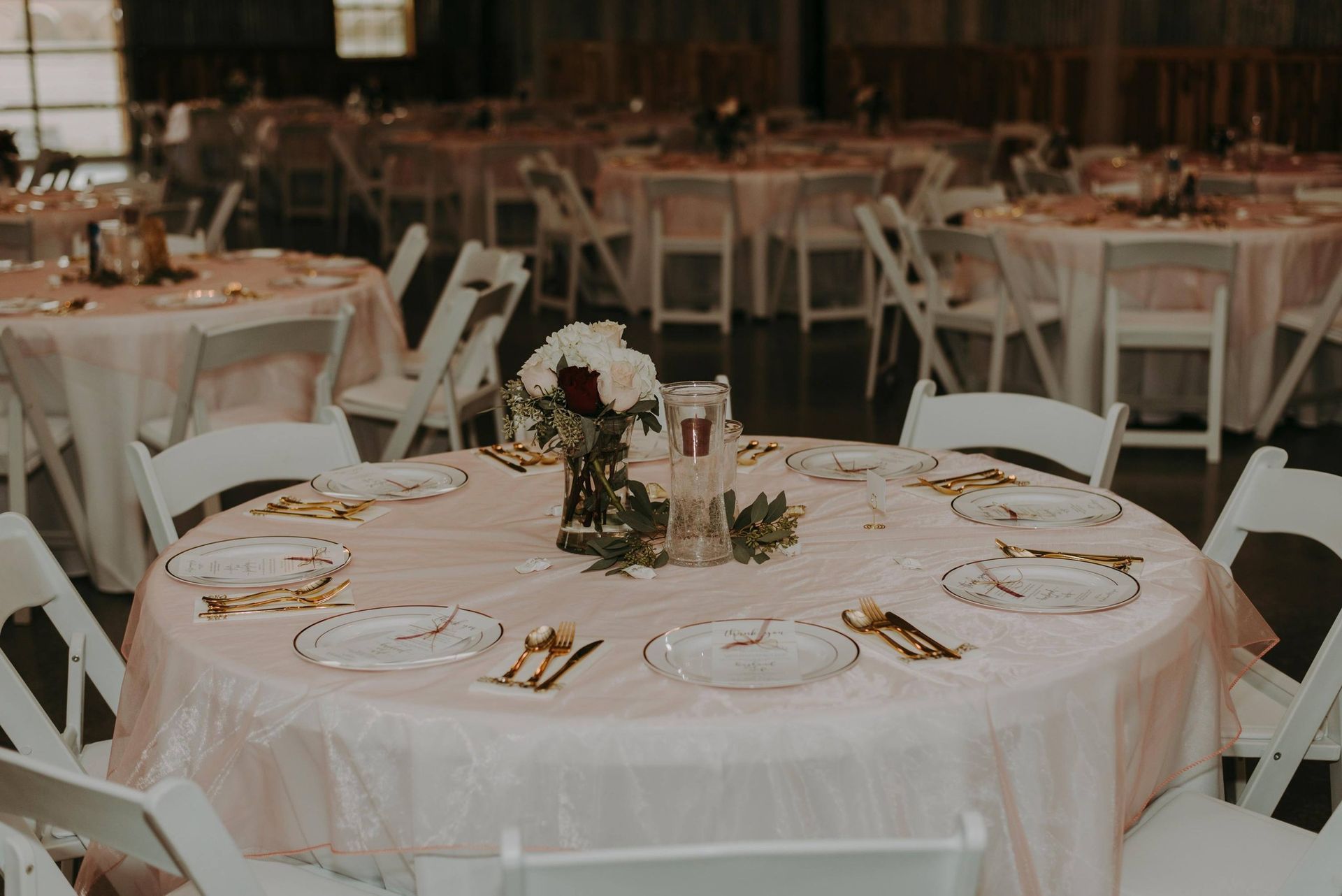 Round tables set for a wedding reception with white chairs and pink tablecloths. Centerpieces of flowers and candles.