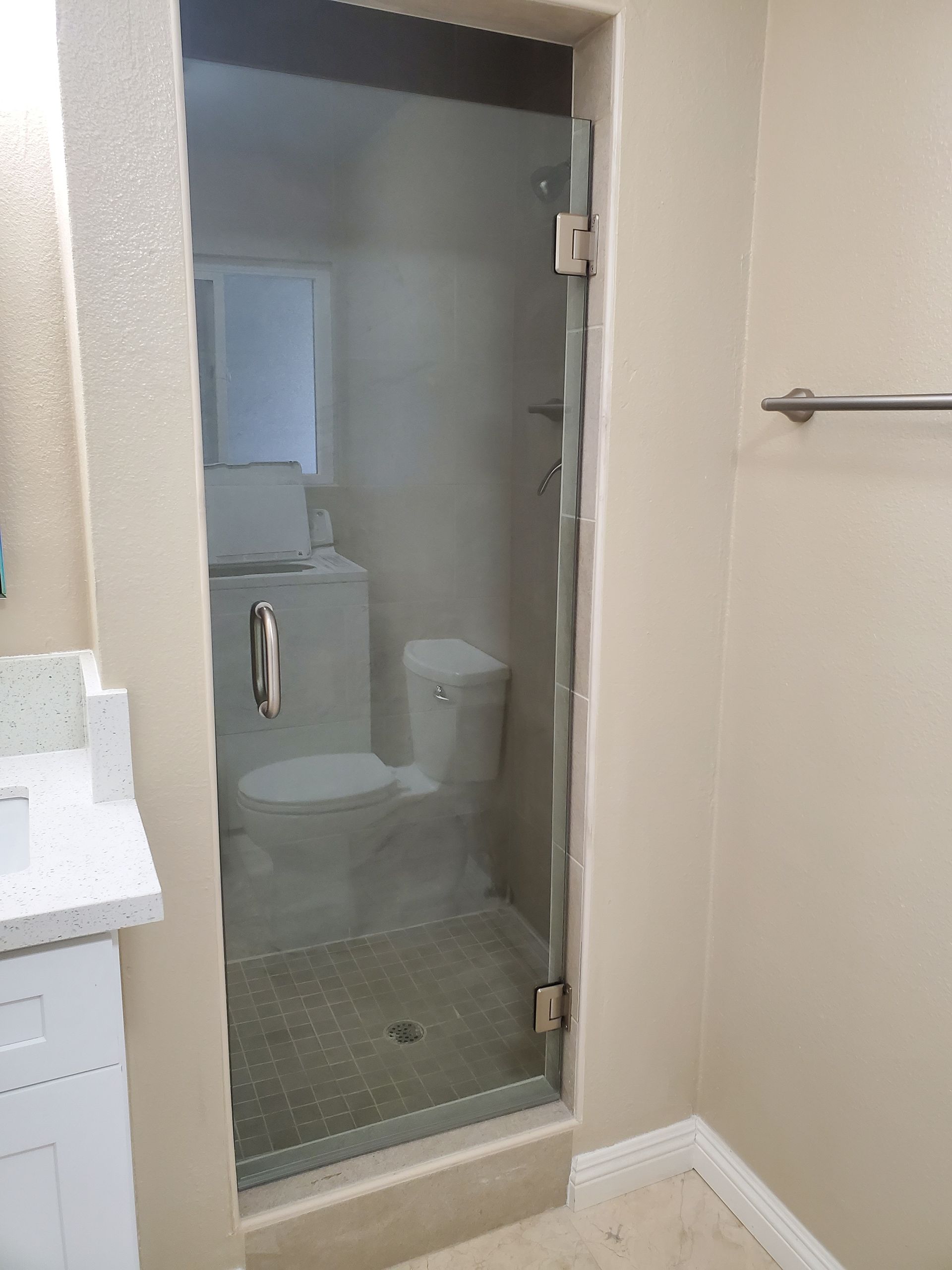 A modern bathroom features a glass shower door with a silver handle and hinges, set against light beige walls.