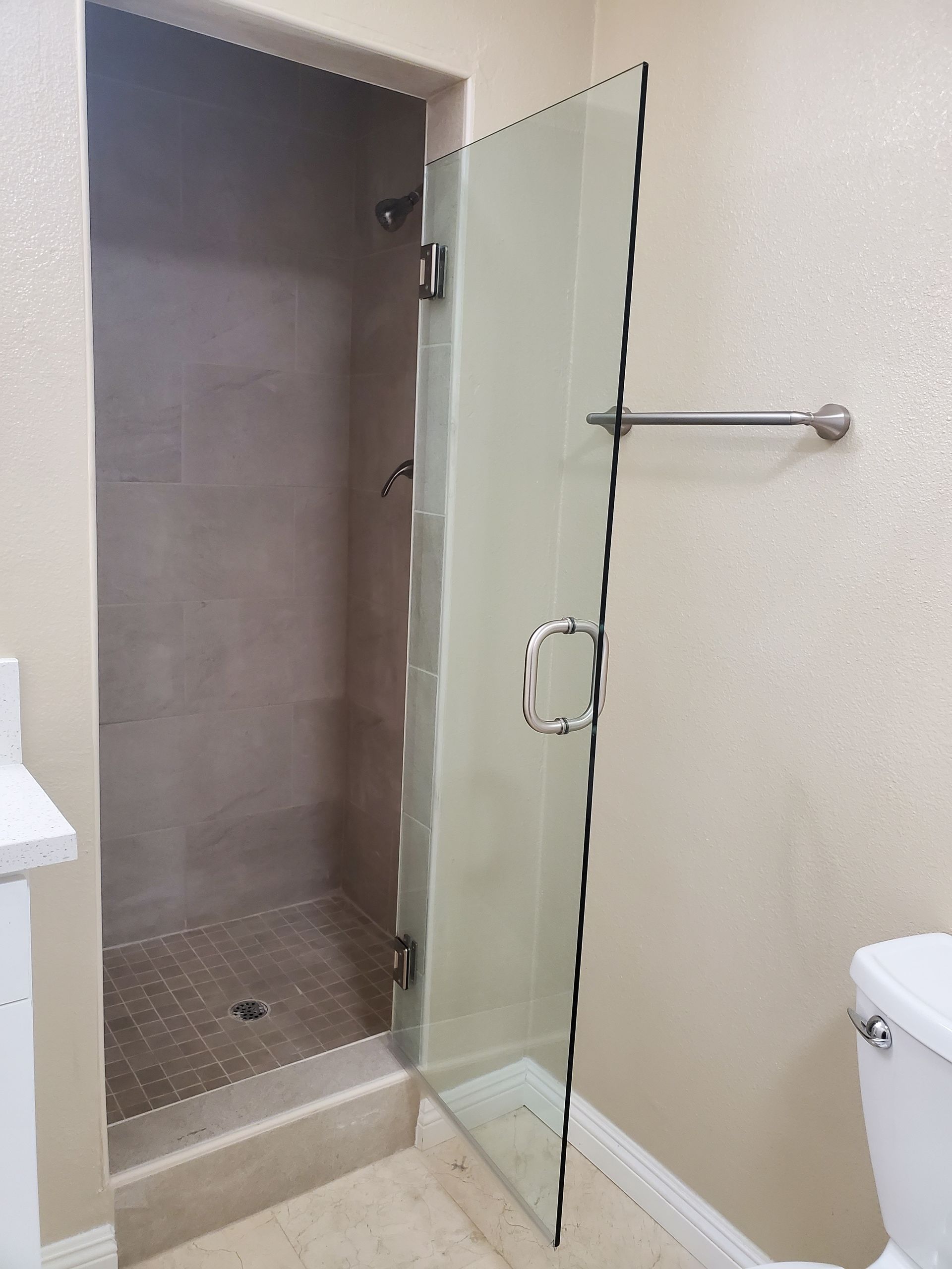 A tiled walk-in shower with a glass door in a neutral-toned bathroom with a towel bar and toilet nearby.