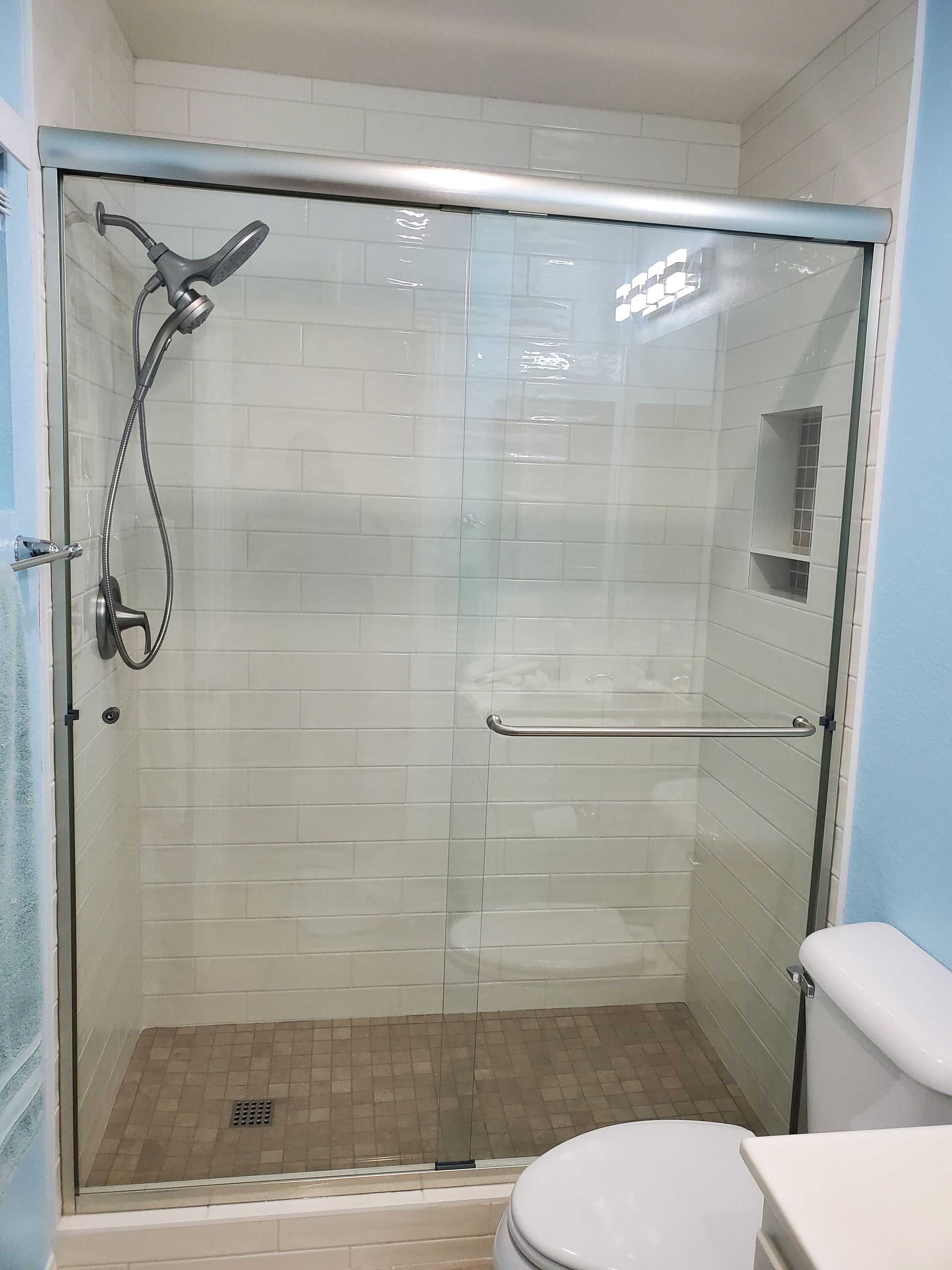 A walk-in shower with white tiled walls, a glass sliding door, handheld shower head, and a recessed storage niche.