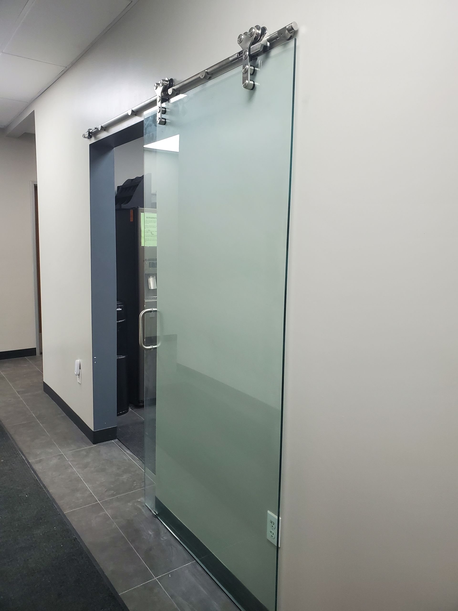 Frosted glass sliding door hanging from a silver track in a modern office hallway.