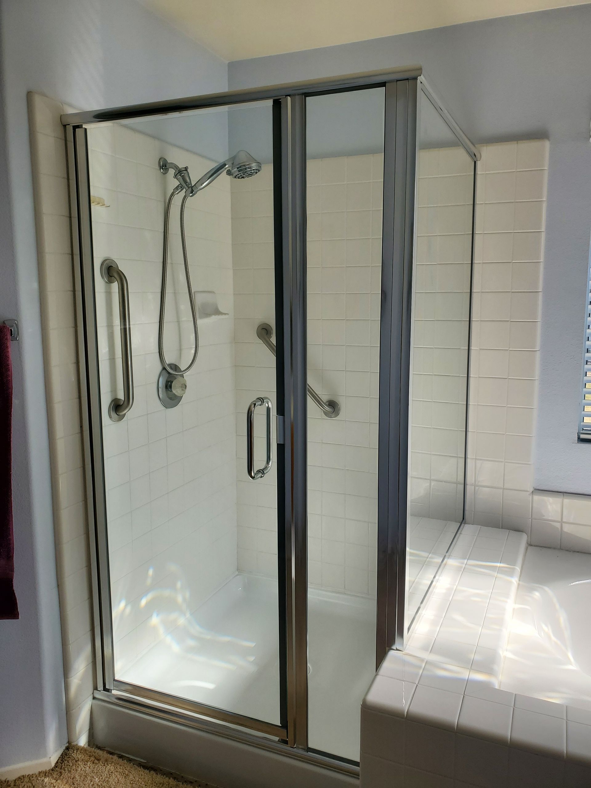 A corner shower stall with a glass door, chrome fixtures, and white tile walls featuring two safety grab bars.
