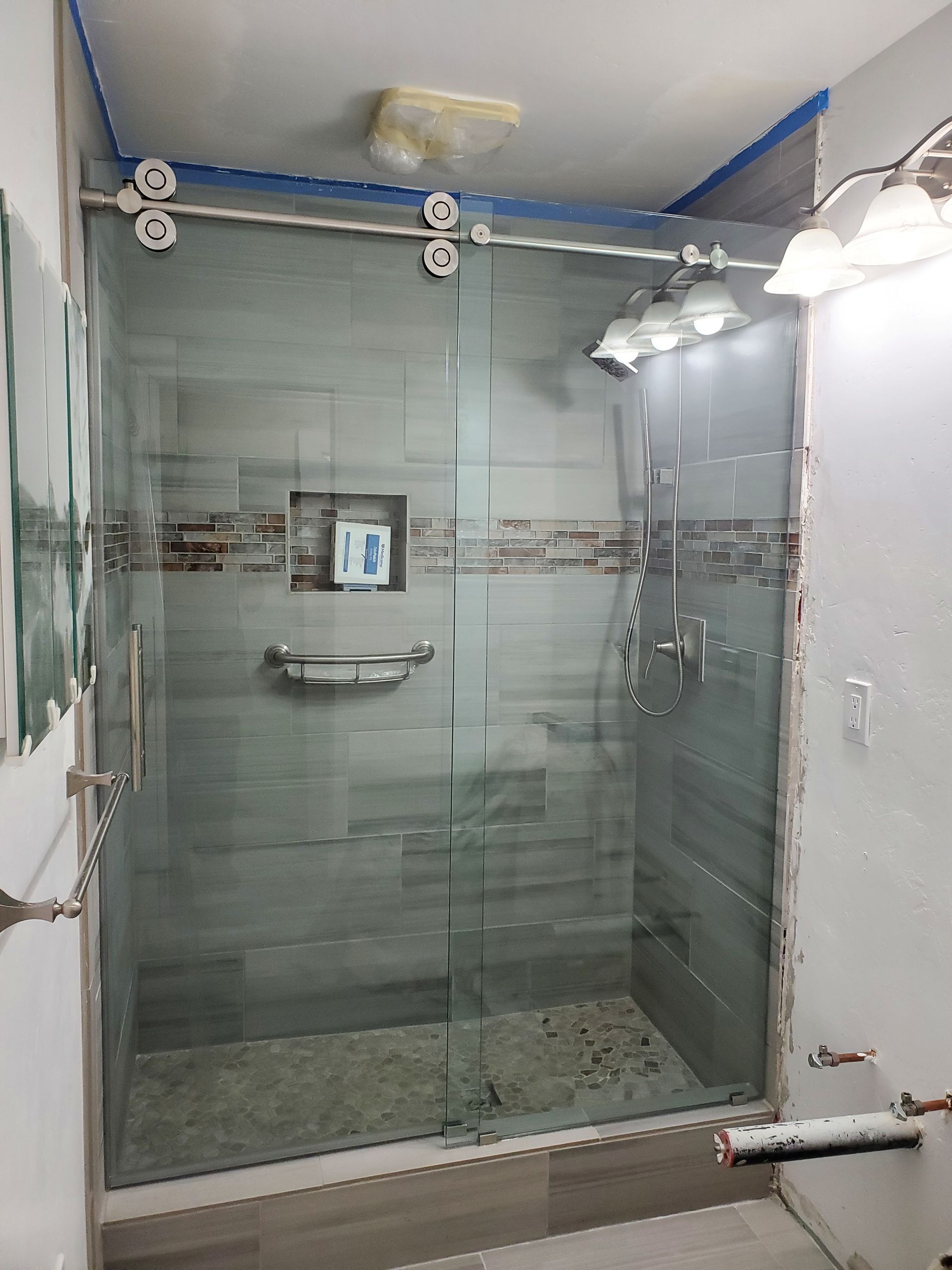 A modern glass-enclosed shower with gray tiled walls, a small recessed shelf, and pebble-style flooring.