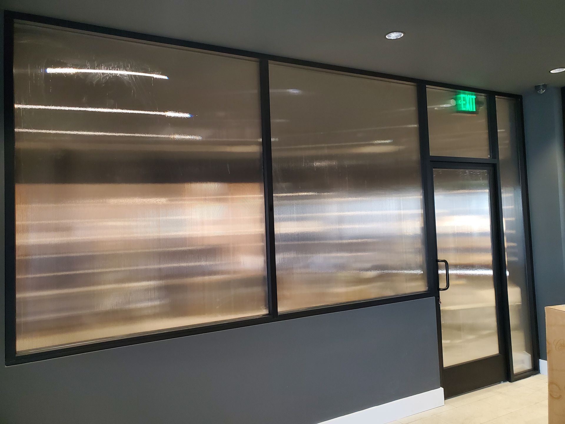 A glass-walled office entrance with black framing, a bright green illuminated exit sign, and gray walls.