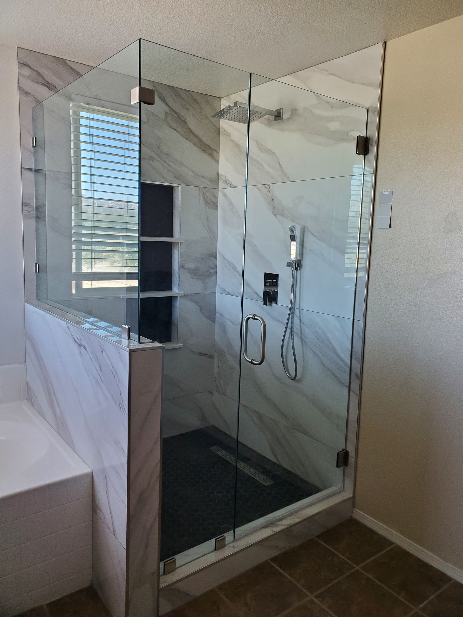 A frameless glass shower enclosure with marble tile walls, a dark floor, and chrome fixtures in a bathroom.