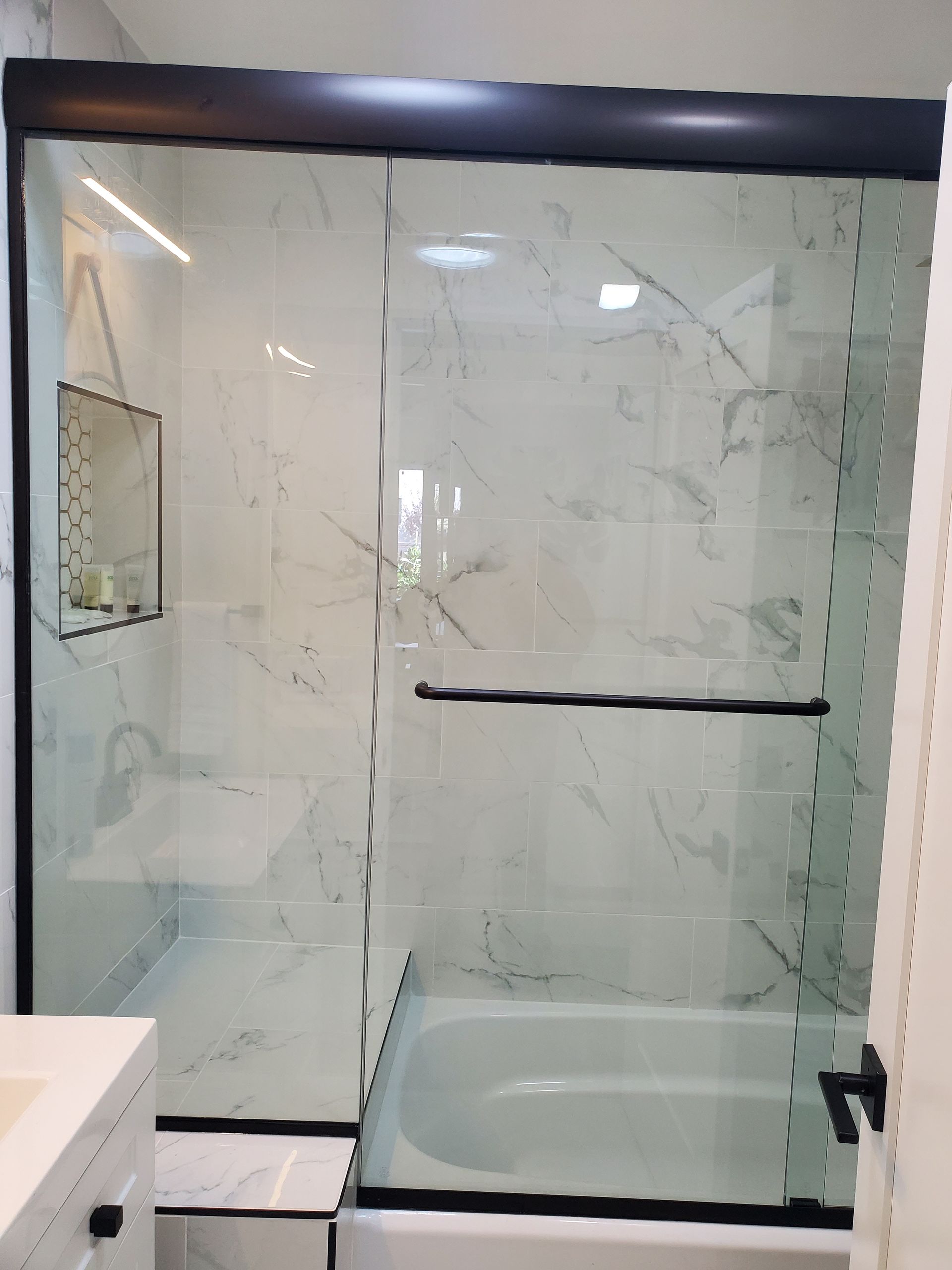 A bathroom features a bathtub with a sliding glass door trimmed in black, set against marble-patterned tile walls.