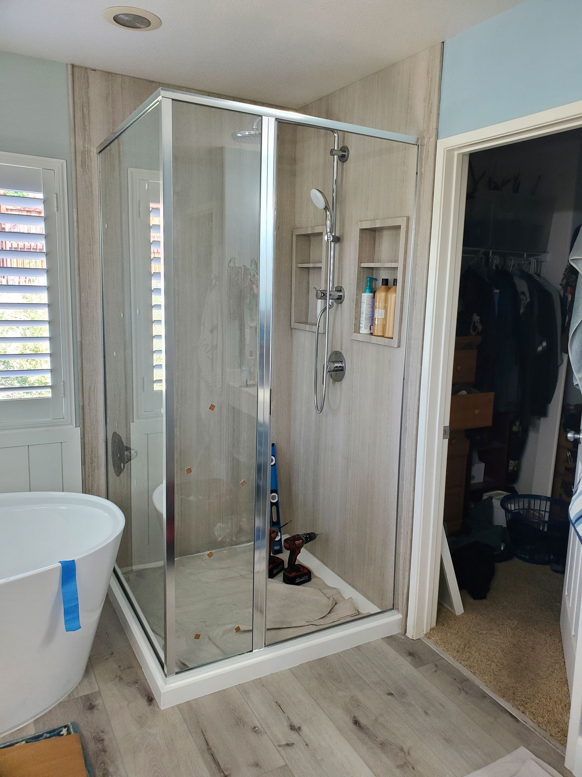 A corner shower stall with a glass door and wood-look panels, positioned next to a white bathtub and an open closet.