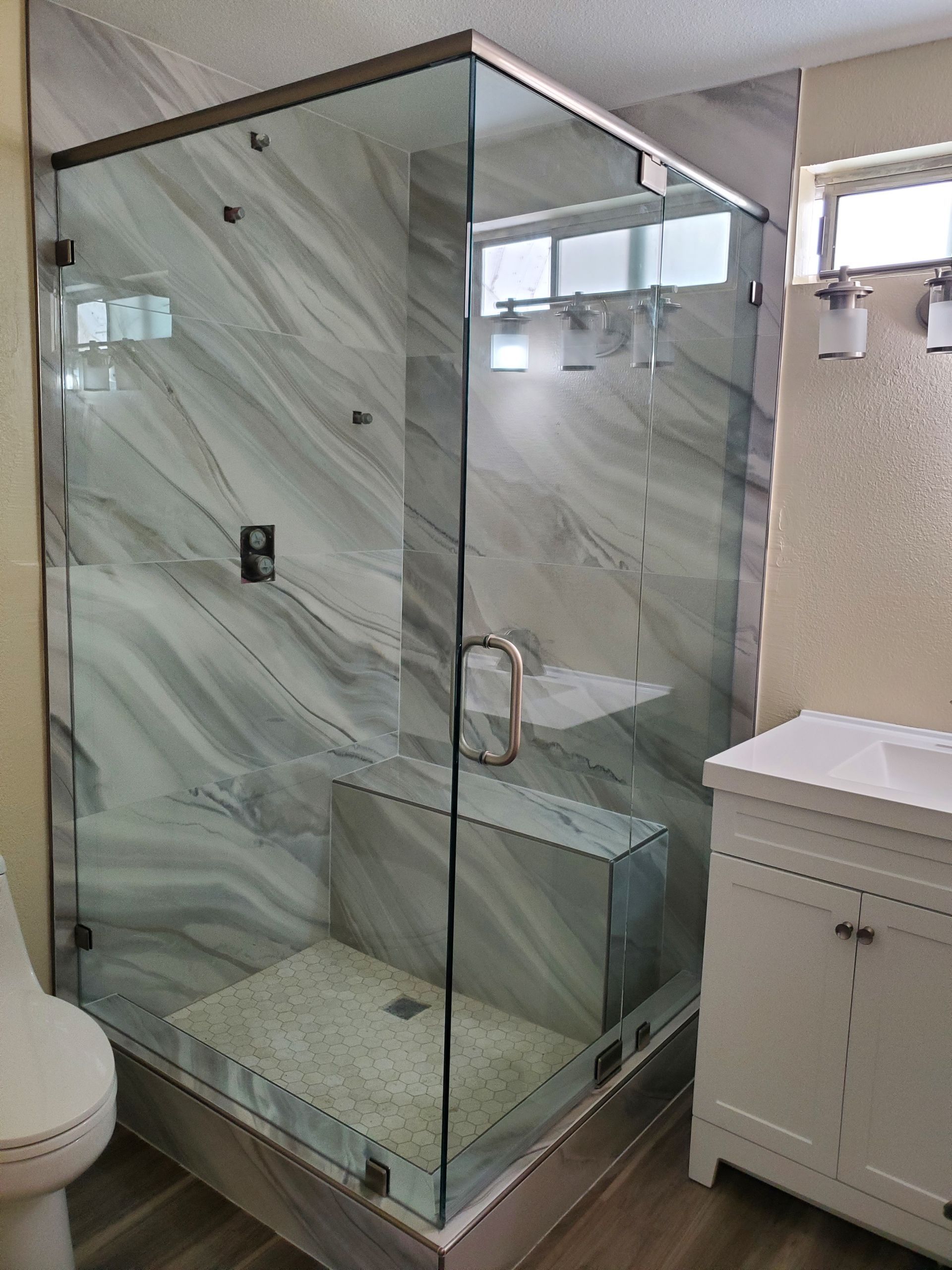 A corner walk-in glass shower with marble-patterned walls and a built-in bench seat next to a white bathroom vanity.