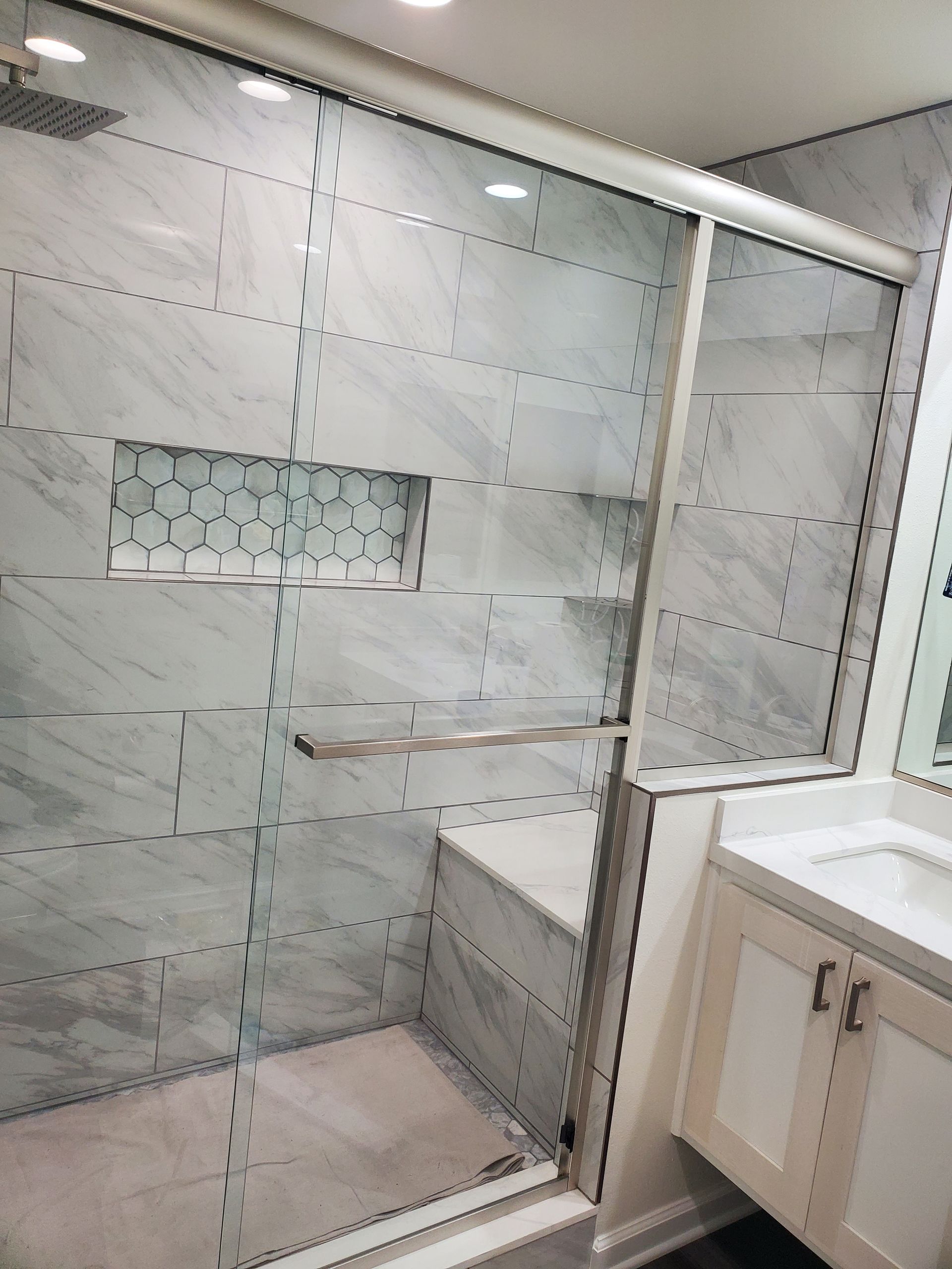 A modern shower with marble-look wall tiles, a built-in bench, a recessed shelf with hexagonal tiles, and glass doors.