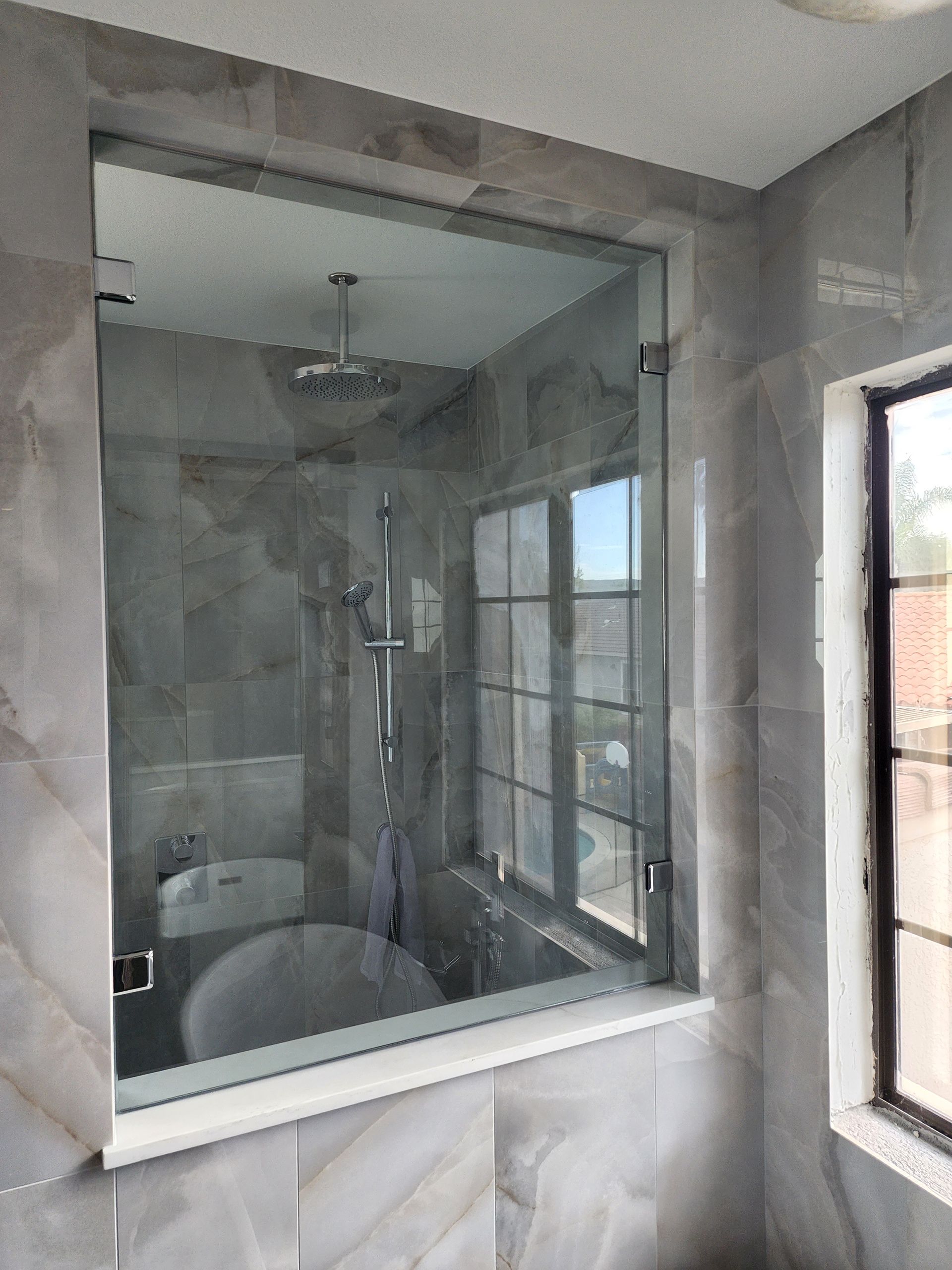 A glass shower door set against gray-toned tiled walls, with a window visible in the background and through the glass.