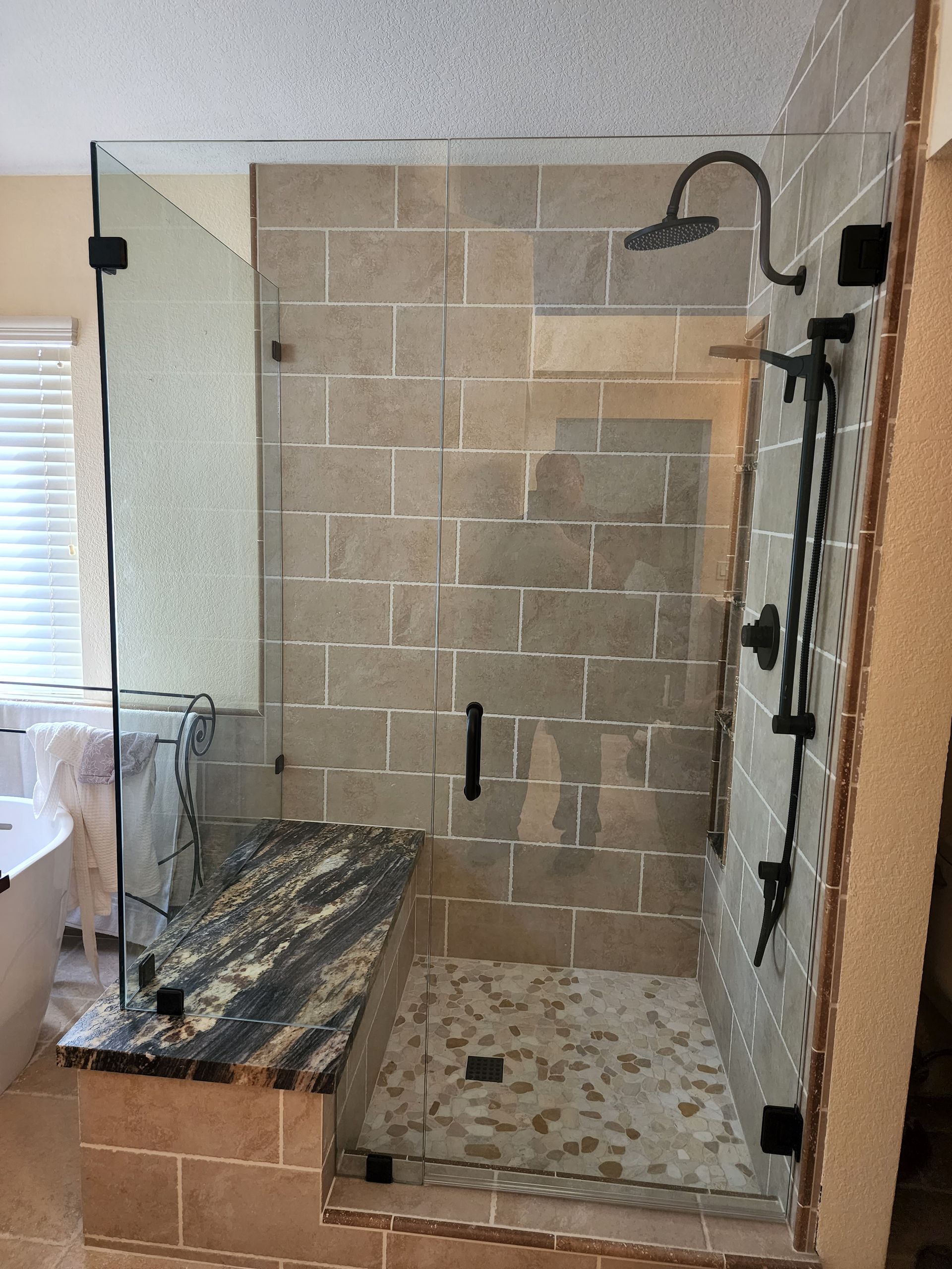 A glass-enclosed shower with beige stone-look tile walls, a dark granite bench, and black fixtures.