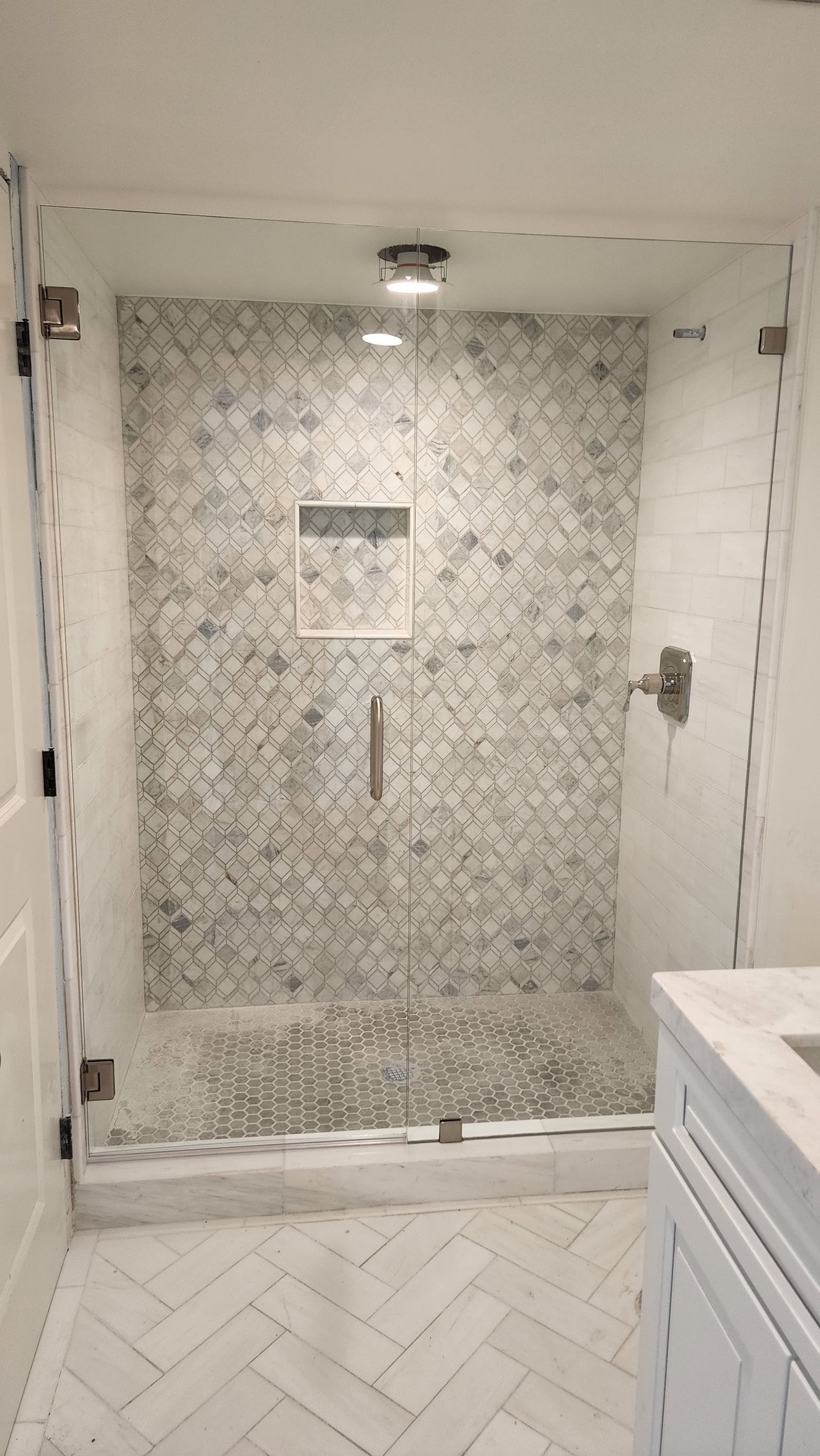A walk-in shower with white and grey patterned tiles, a marble-look floor, and a glass door.