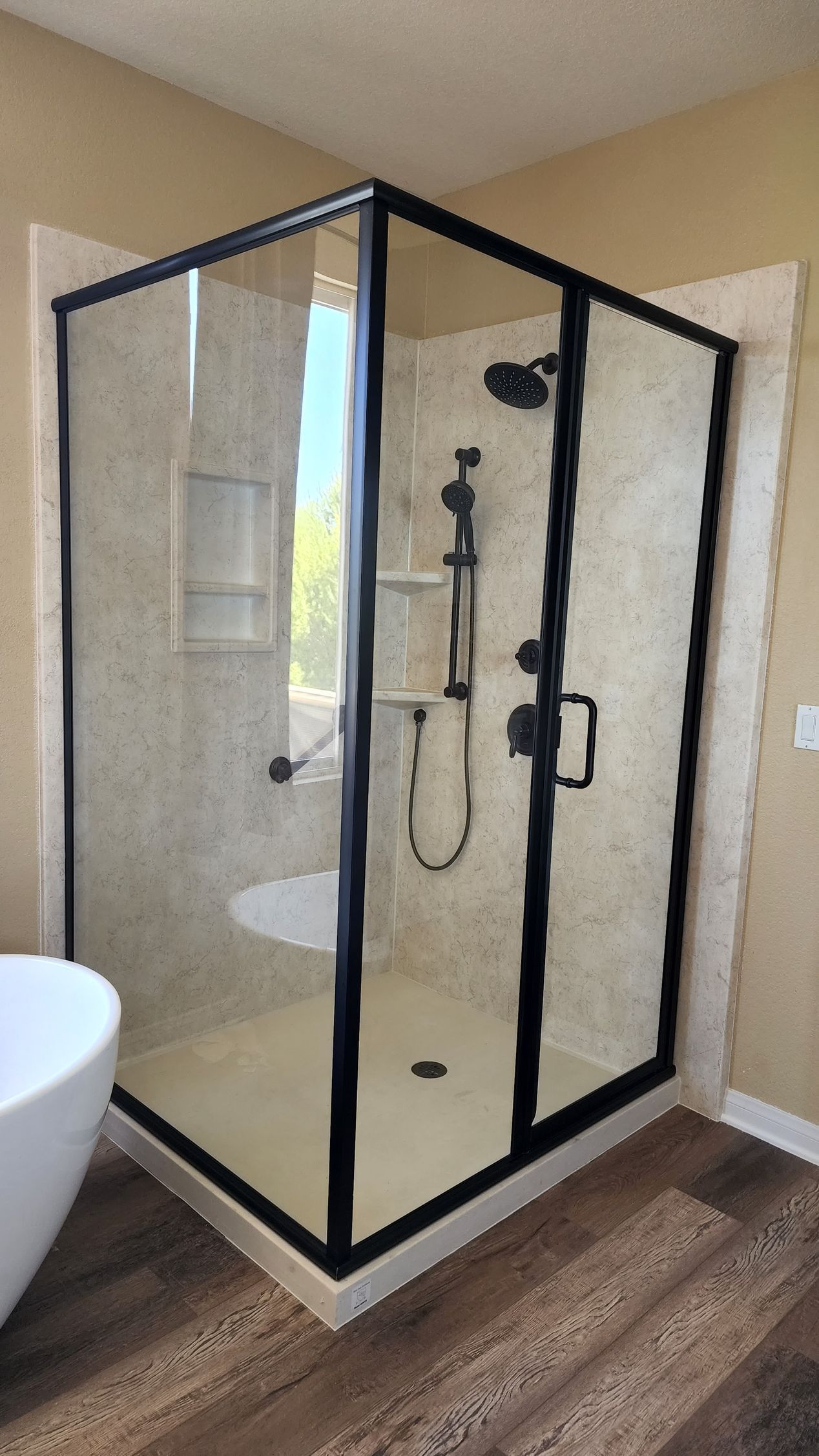A corner shower stall with a black frame, glass door, and cream-colored tiled walls in a bathroom with wood-look flooring.