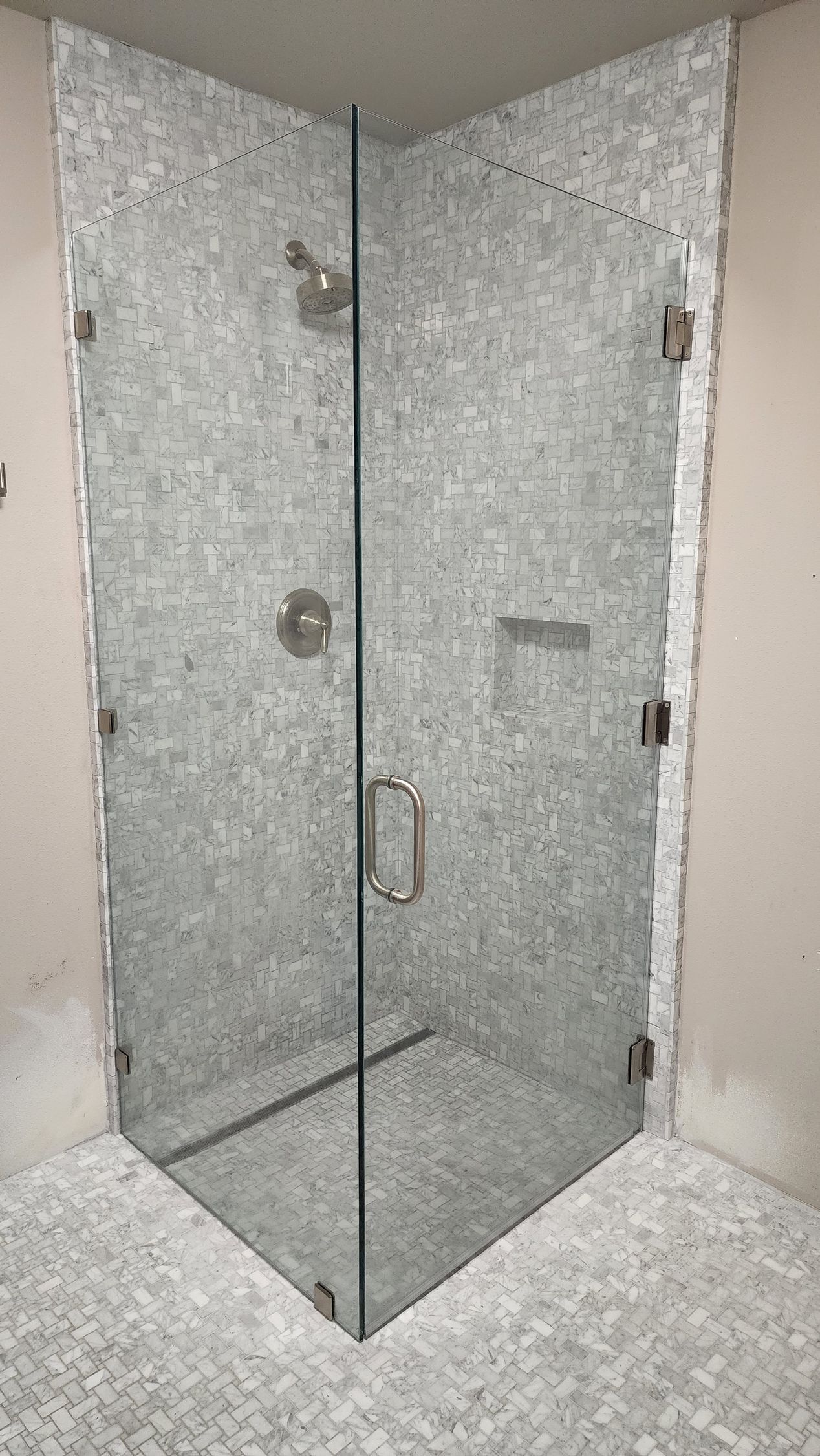 A corner glass shower enclosure with chrome hardware and light-colored mosaic tile walls and floor in an unfinished room.