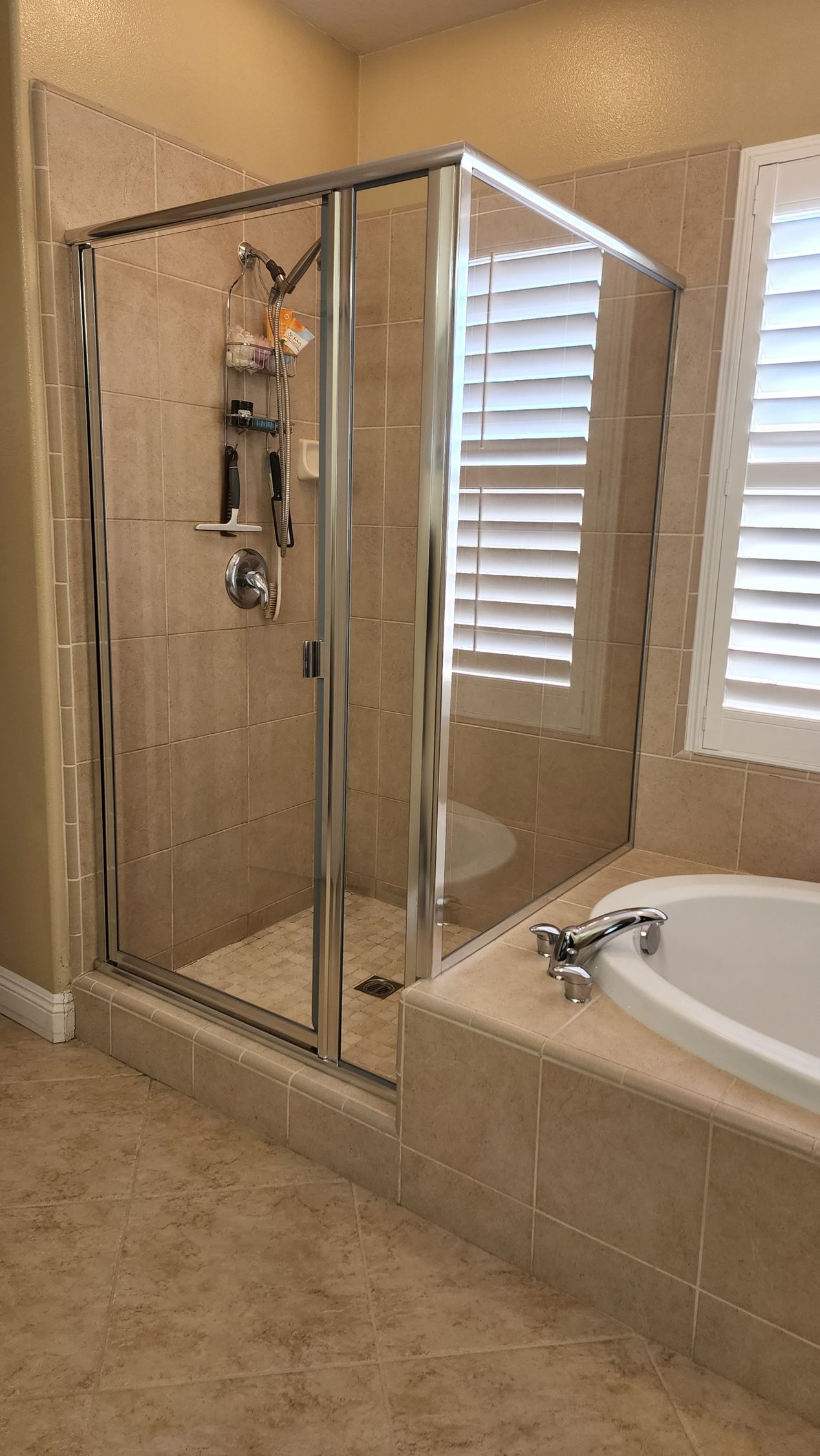 A glass-enclosed shower with beige tile walls next to a bathtub and window with white shutters in a bathroom.