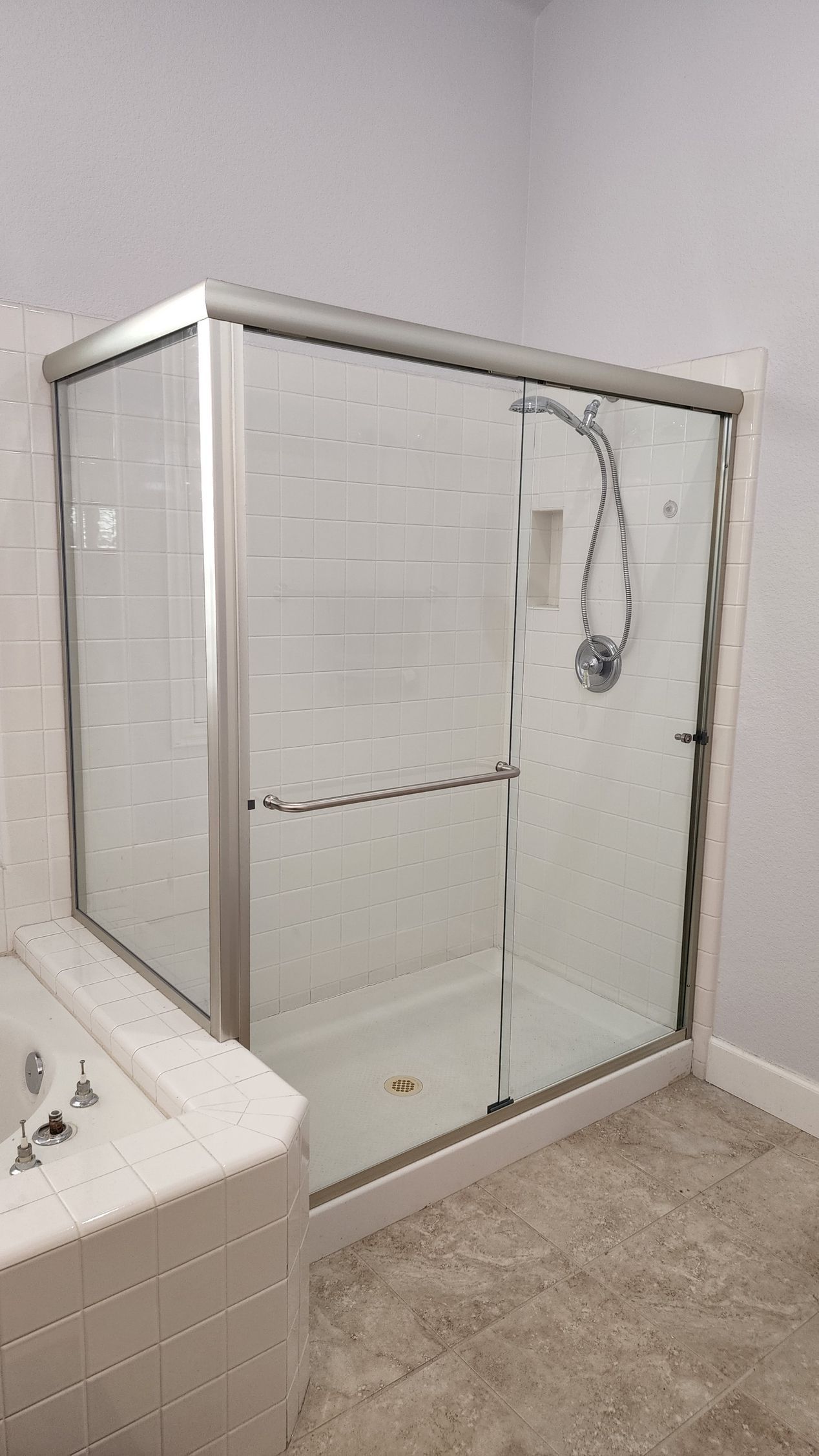 A glass-enclosed shower stall with a silver frame next to a tiled bathtub in a bathroom with beige flooring.