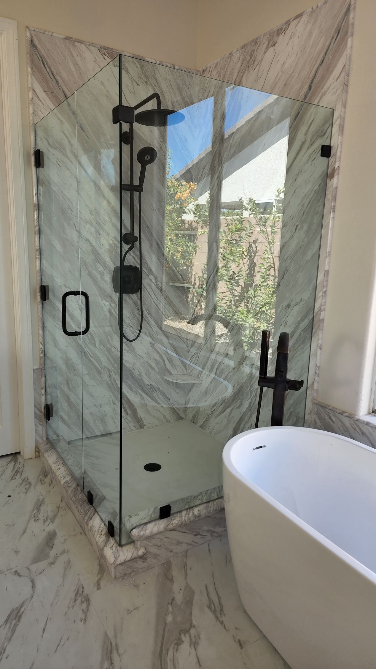 A corner glass shower stall with matte black fixtures sits next to a white freestanding bathtub in a tiled bathroom.