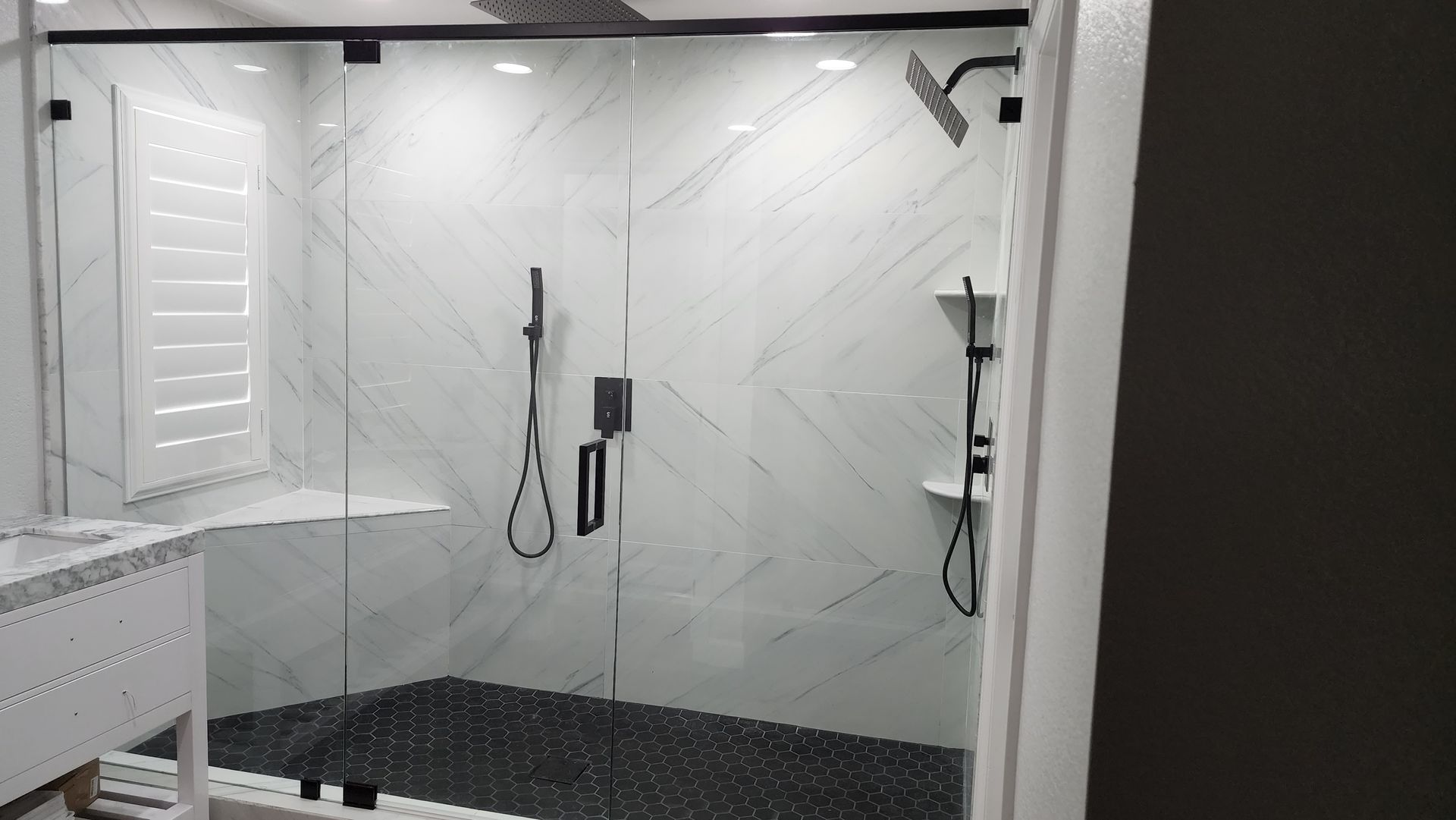 A modern walk-in shower with white marble-patterned tile, black fixtures, glass doors, and a dark mosaic tile floor.
