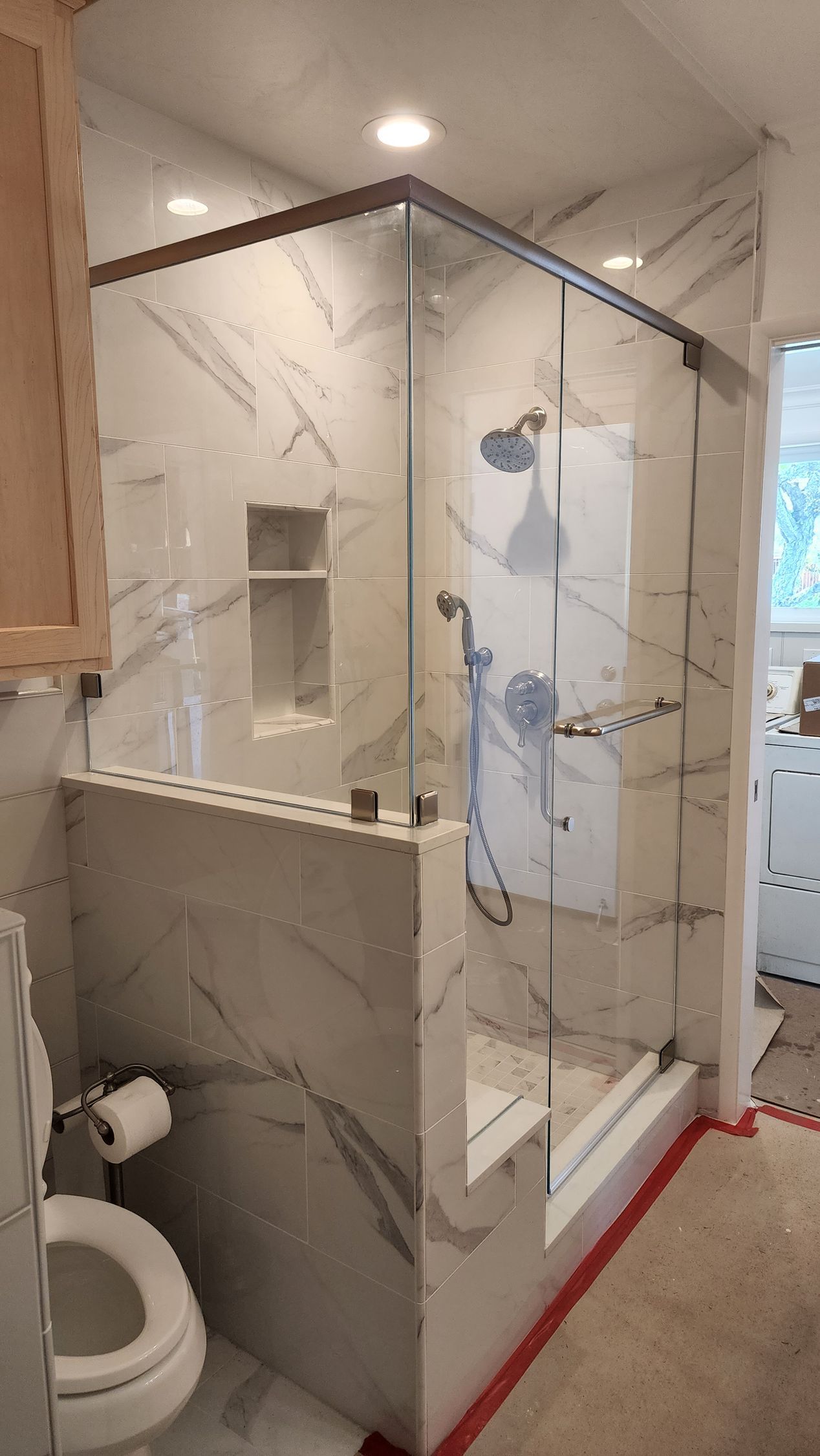 A modern bathroom shower with marble-tiled walls, glass enclosure, built-in shelving, and a toilet nearby.