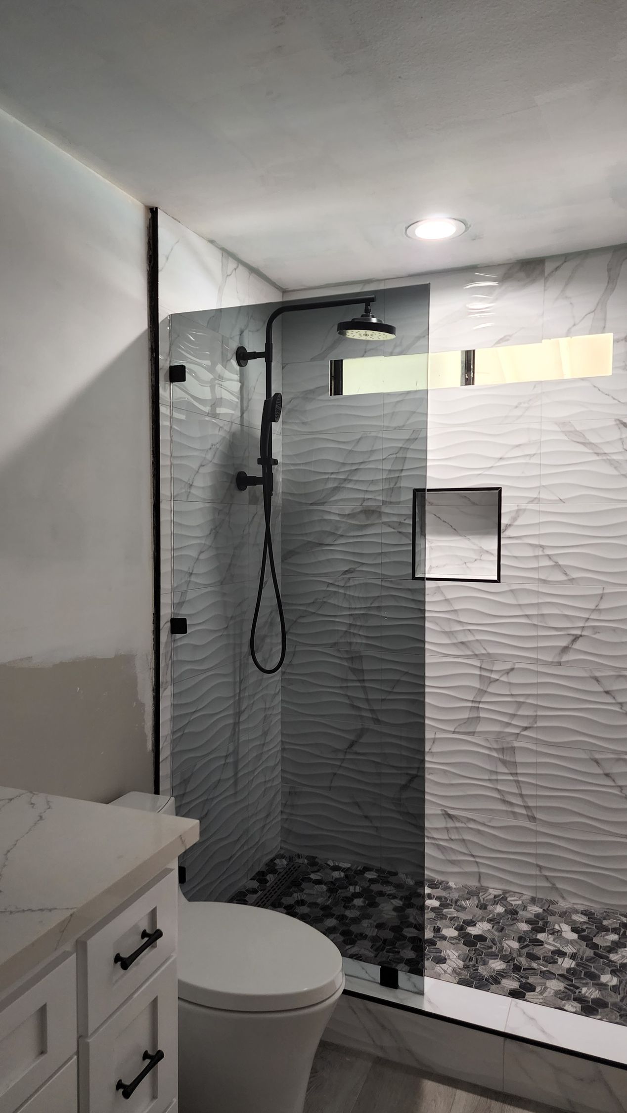 A modern bathroom with a glass-enclosed shower featuring textured white marble walls, black fixtures, and a pebble floor.