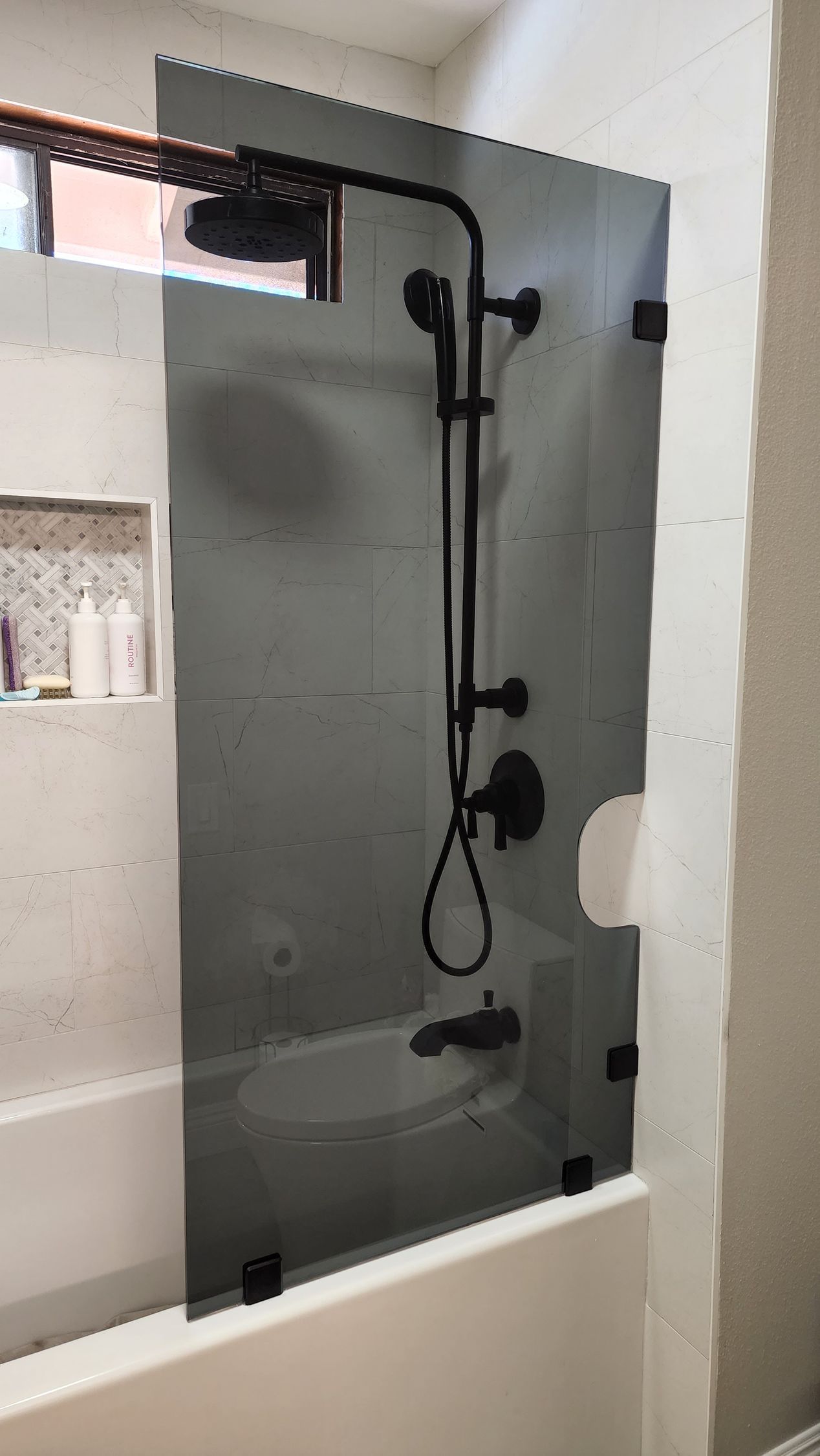 A matte black shower fixture and a tinted glass panel mounted above a white bathtub in a tiled bathroom.