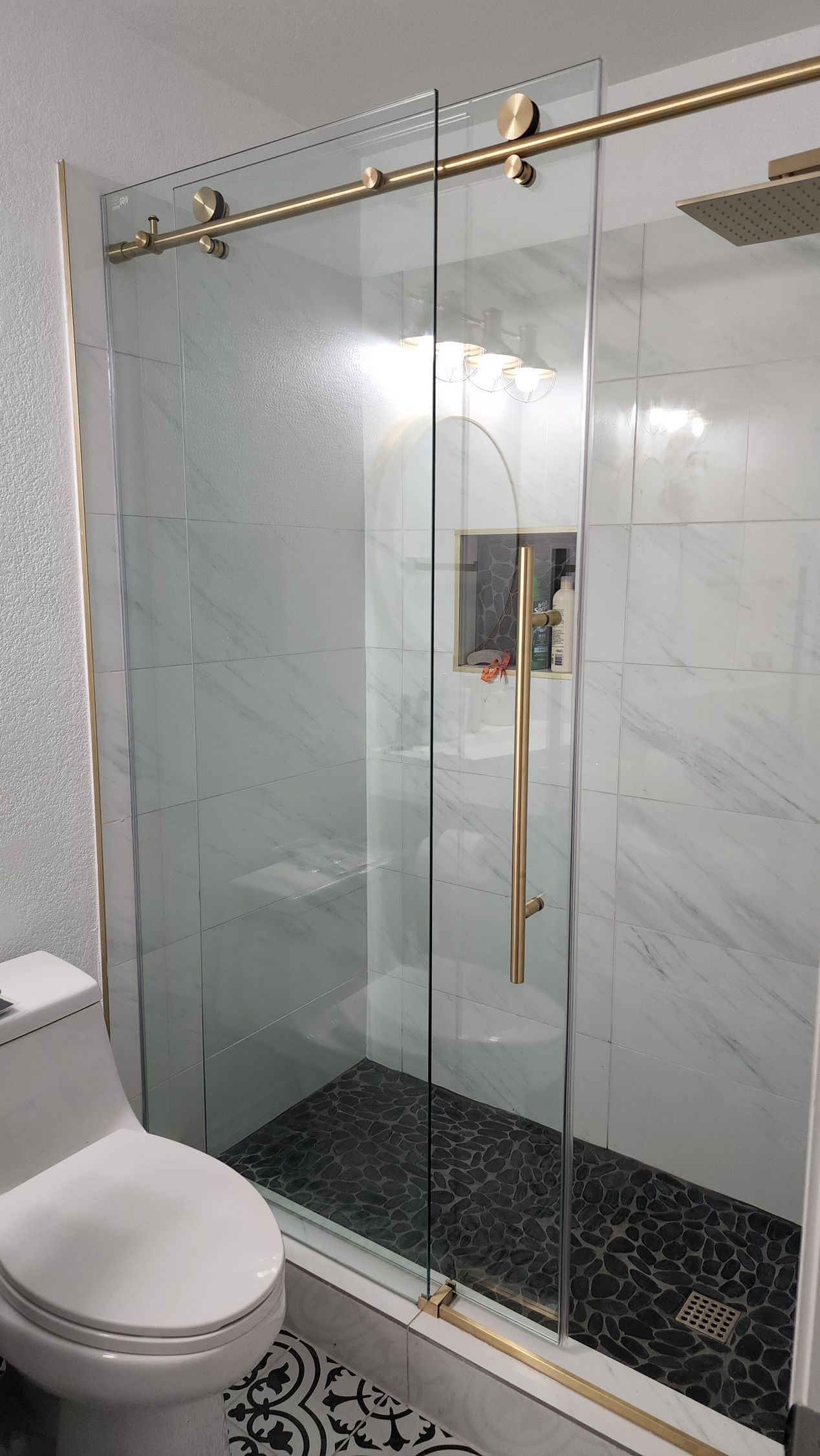 A modern walk-in shower with gold hardware, sliding glass doors, marble-patterned walls, and a black stone floor.