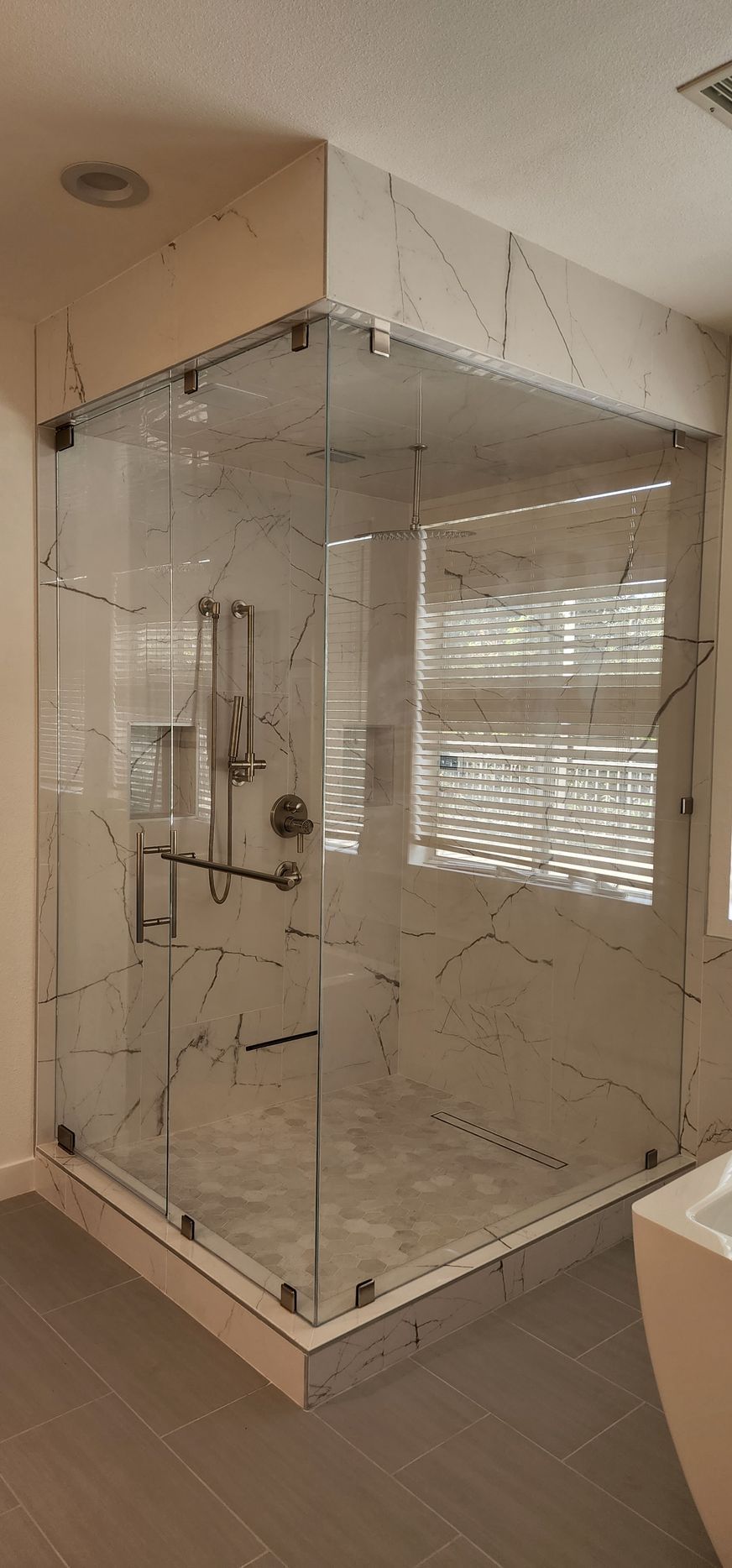 A corner glass shower enclosure with marble-patterned tile walls and floor, featuring brushed metal hardware.