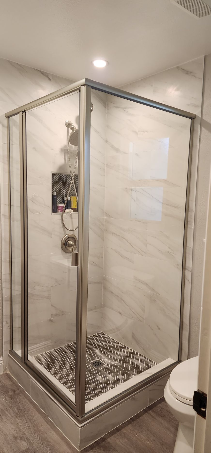 A square shower stall with silver framing, white marble-patterned tiled walls, and a grey pebble-stone floor.