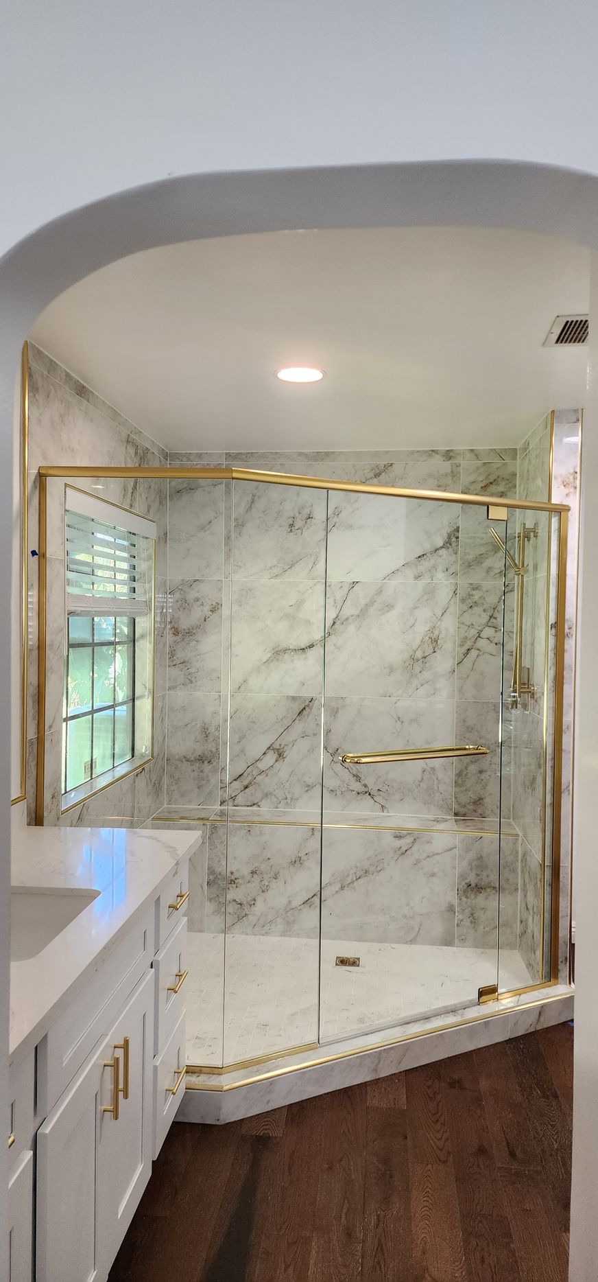 A bathroom featuring a corner shower with glass doors, gold hardware, and marble tile walls next to a white vanity.