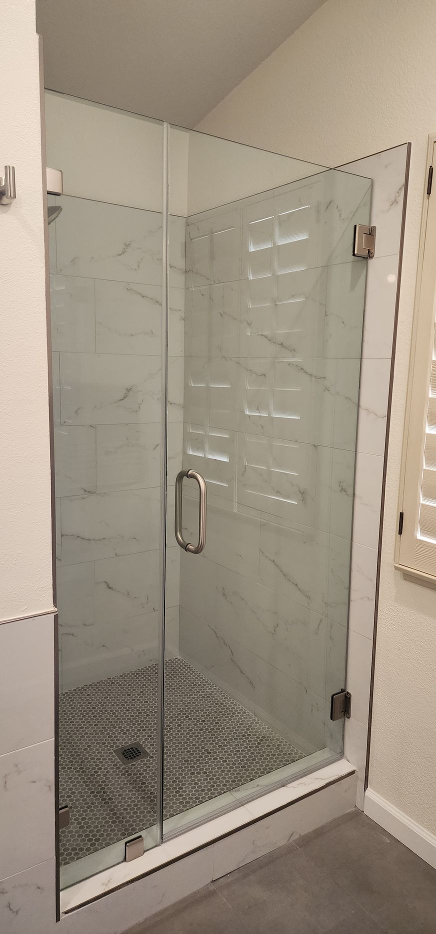 A glass shower door with a silver handle and hinges, installed in a bathroom with marbled tiled walls and a mosaic floor.