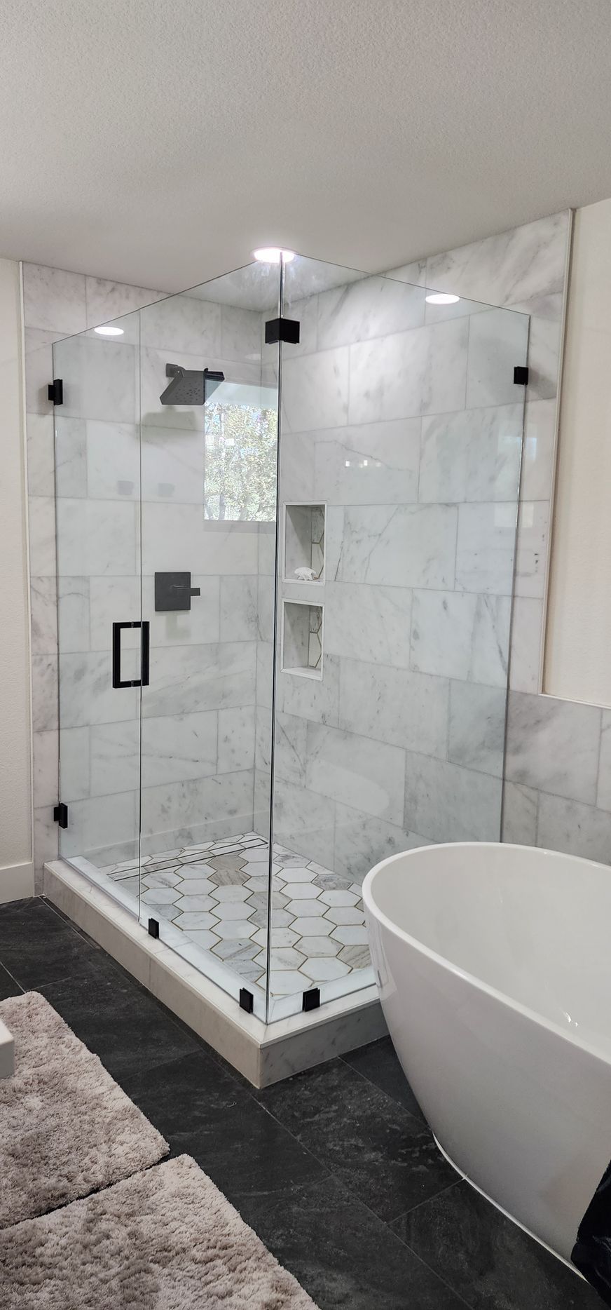 A corner glass walk-in shower with white marble-look tiles, black hardware, and a hexagonal tile floor next to a bathtub.