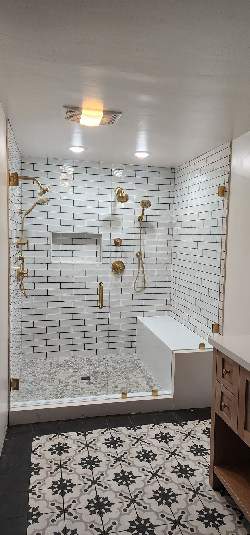 A modern shower with white brick-style wall tiles, patterned floor tile, gold hardware, and a built-in white bench.