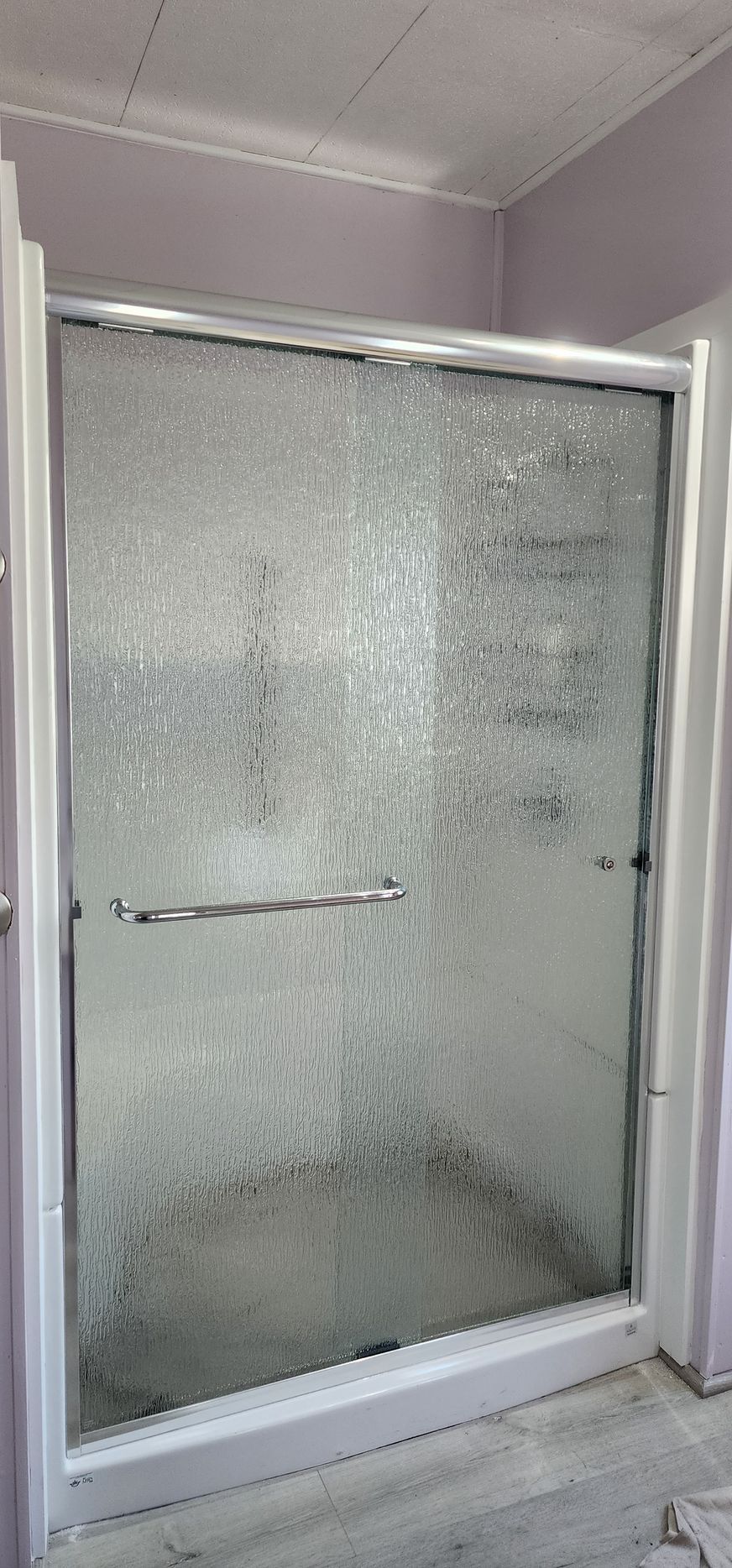 A framed glass shower door with a textured, pebble-like finish and a chrome handle, set in a room with light purple walls.