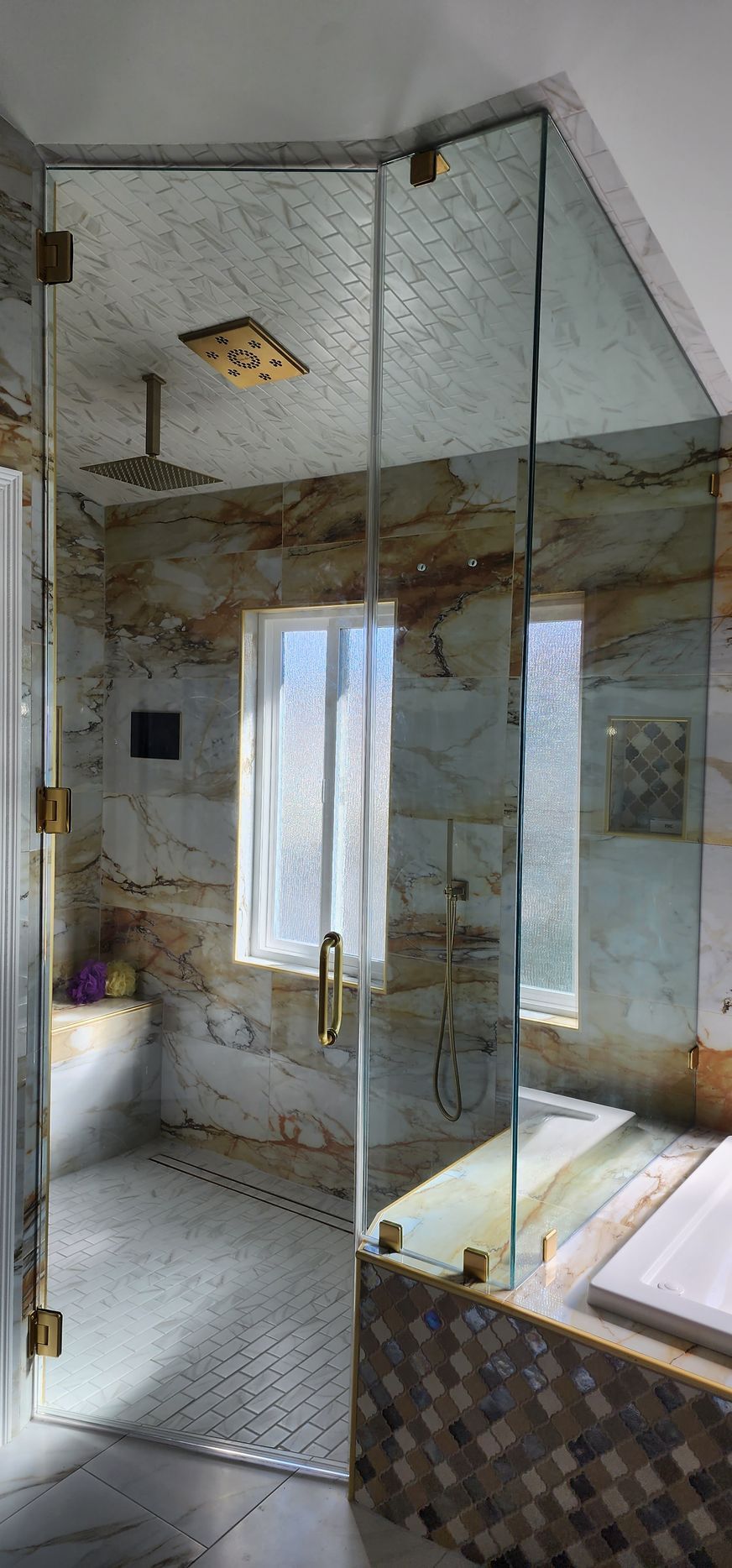 A modern walk-in shower with gold fixtures, marble-patterned walls, and glass doors next to a tiled bathtub.