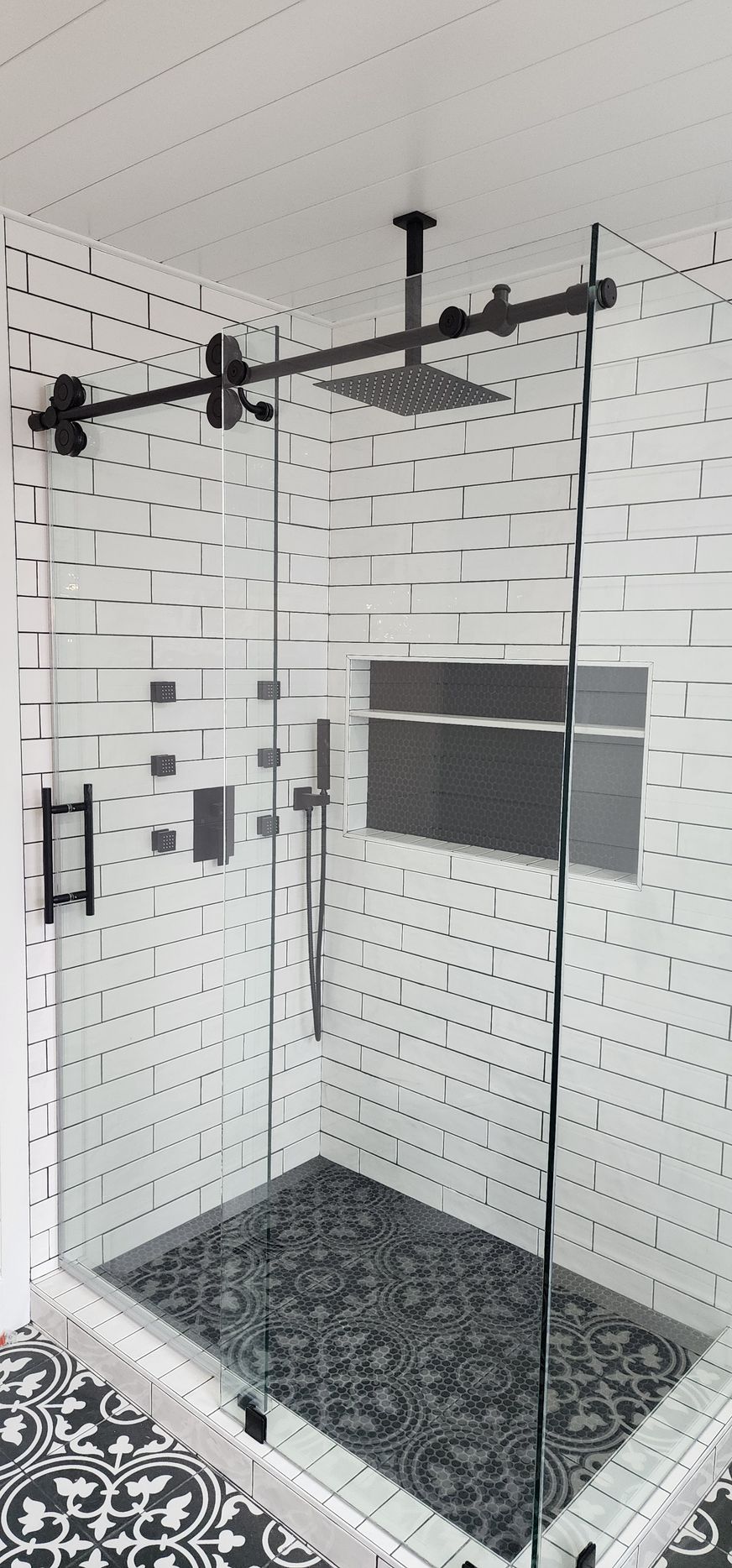 A modern walk-in shower with white subway tiles, a black sliding glass door, a rain showerhead, and a patterned tile floor.