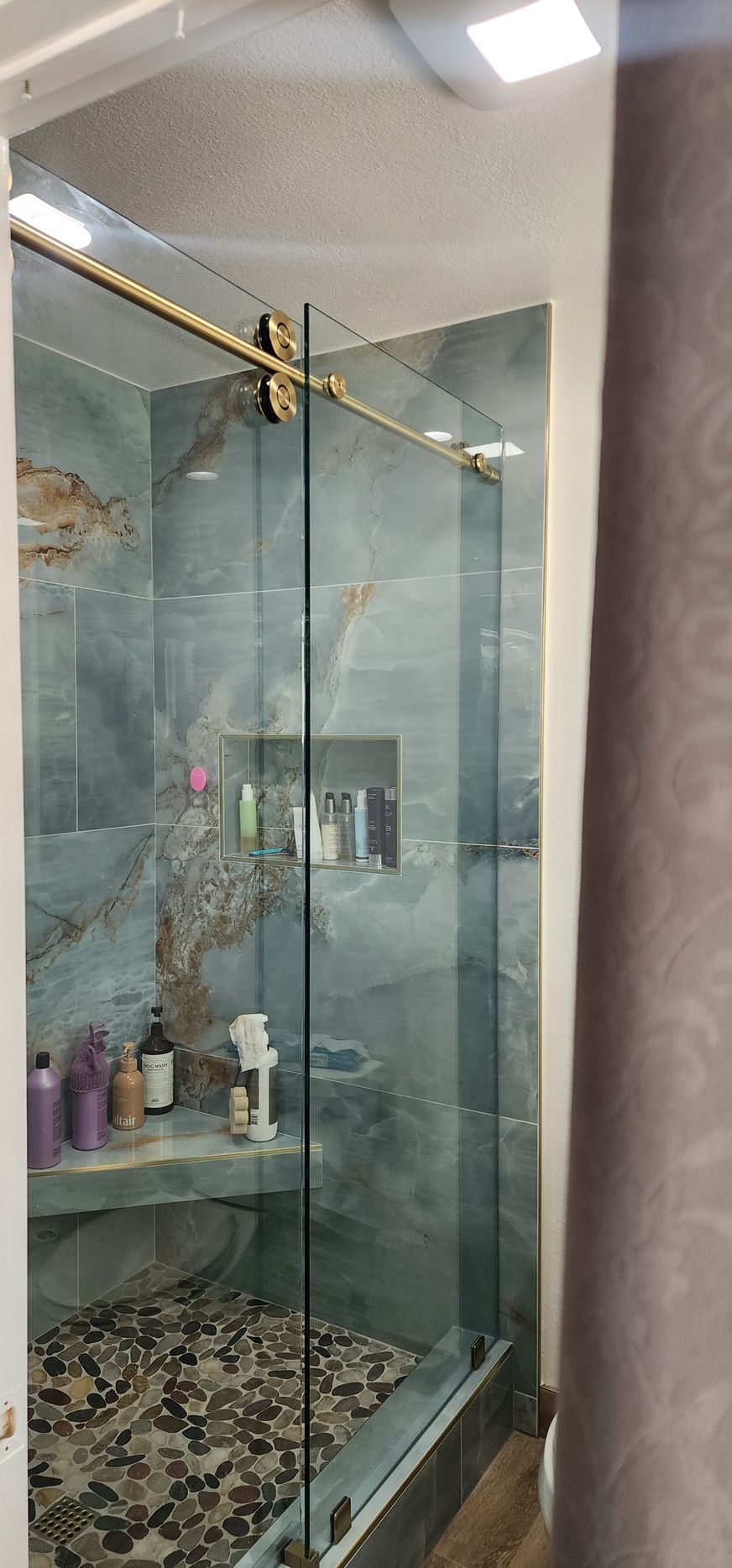 A modern walk-in shower with gray marbled tiles, a pebble floor, a built-in bench, and a glass door with gold hardware.