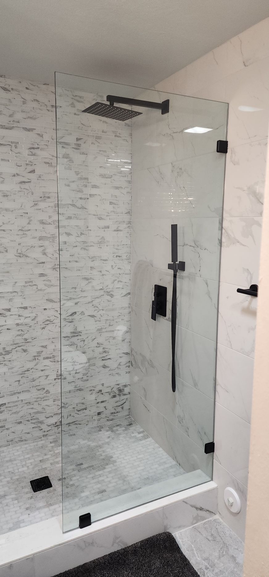A modern glass-enclosed shower with matte black fixtures, a rainfall showerhead, and white marble-tiled walls.
