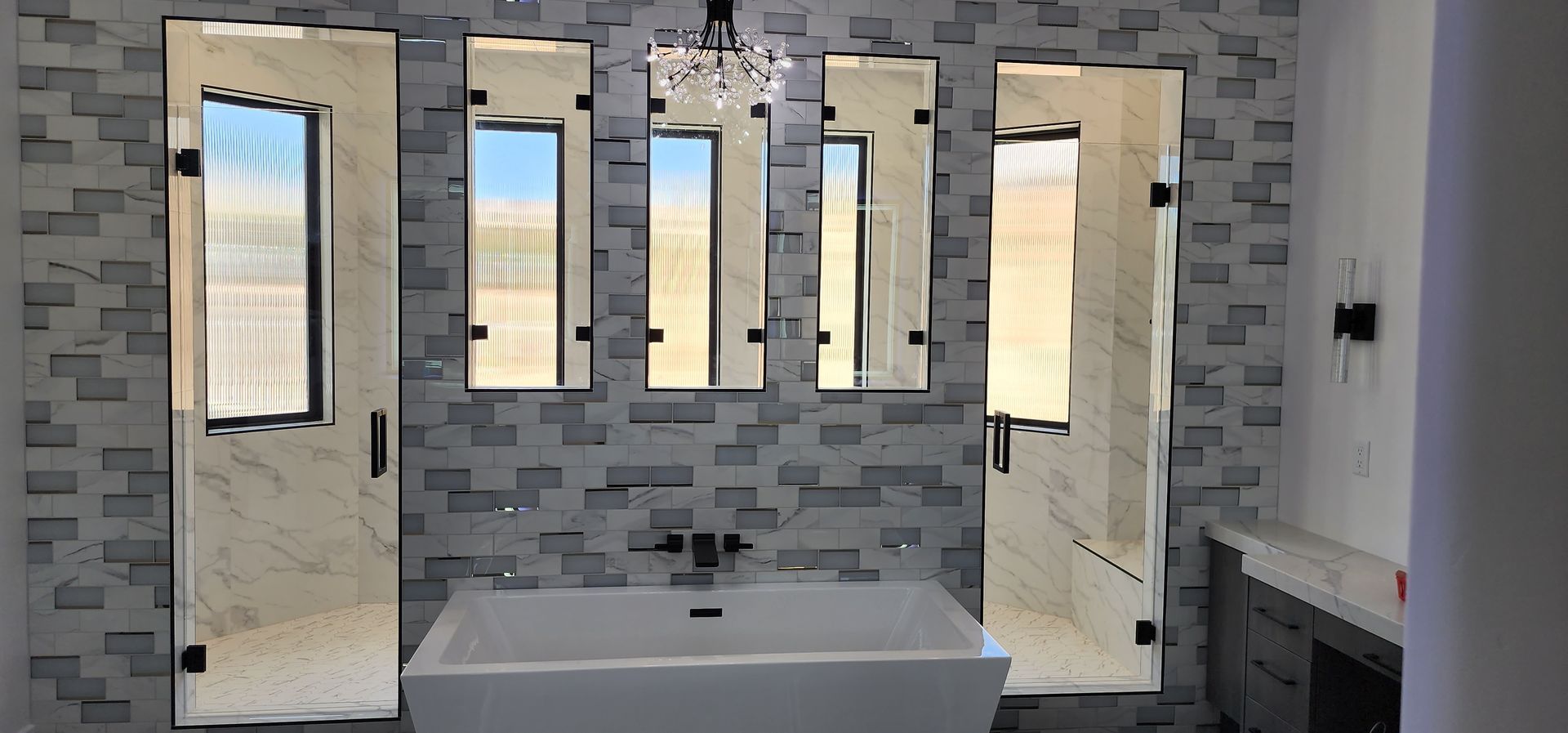 A bathroom with a white freestanding tub, textured gray stone tile walls, three wall mirrors, and glass shower doors.