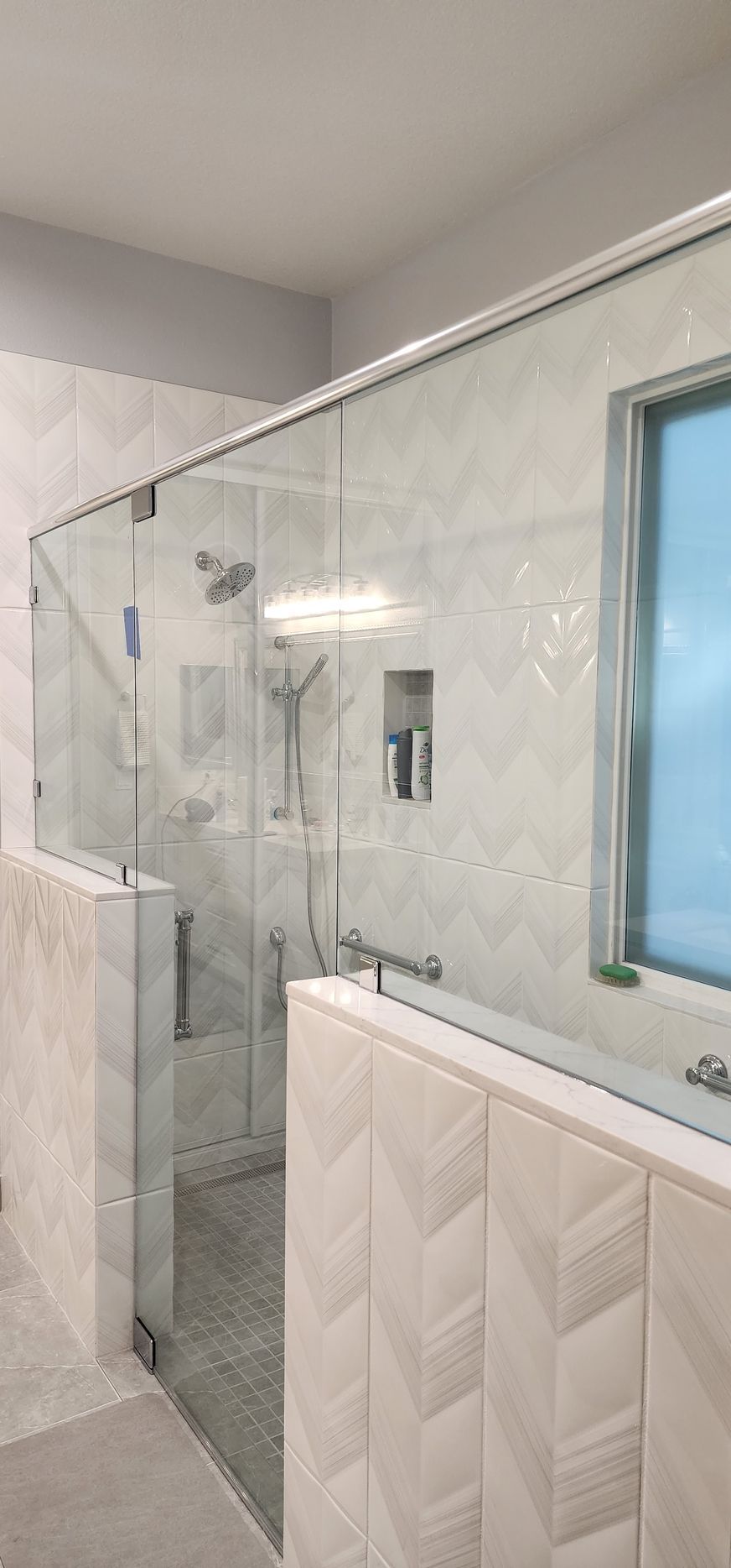 Modern walk-in shower with glass doors and walls, chevron-patterned beige tile, and chrome fixtures.