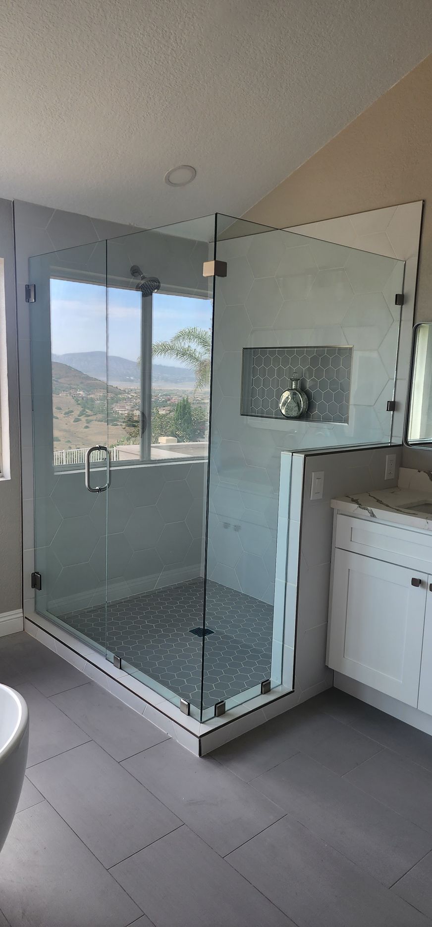 A frameless glass shower enclosure in a bathroom, featuring grey tiled flooring and a view of mountains through a window.
