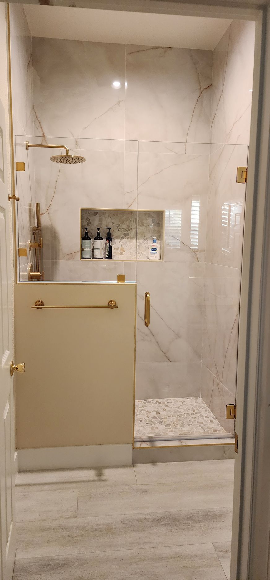 A walk-in shower with marble-style tiles, gold hardware, a glass door, and a recessed niche with toiletries.