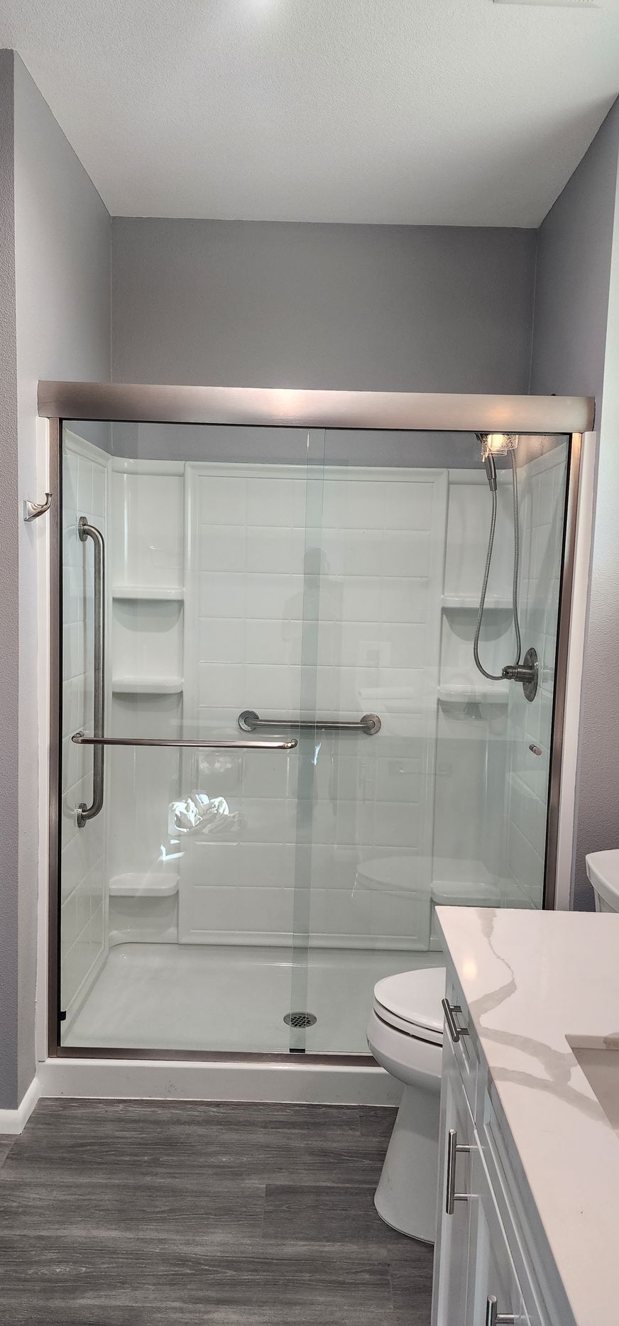 Modern bathroom with a glass-enclosed shower, white tiled walls with integrated shelves, and gray wood-look flooring.