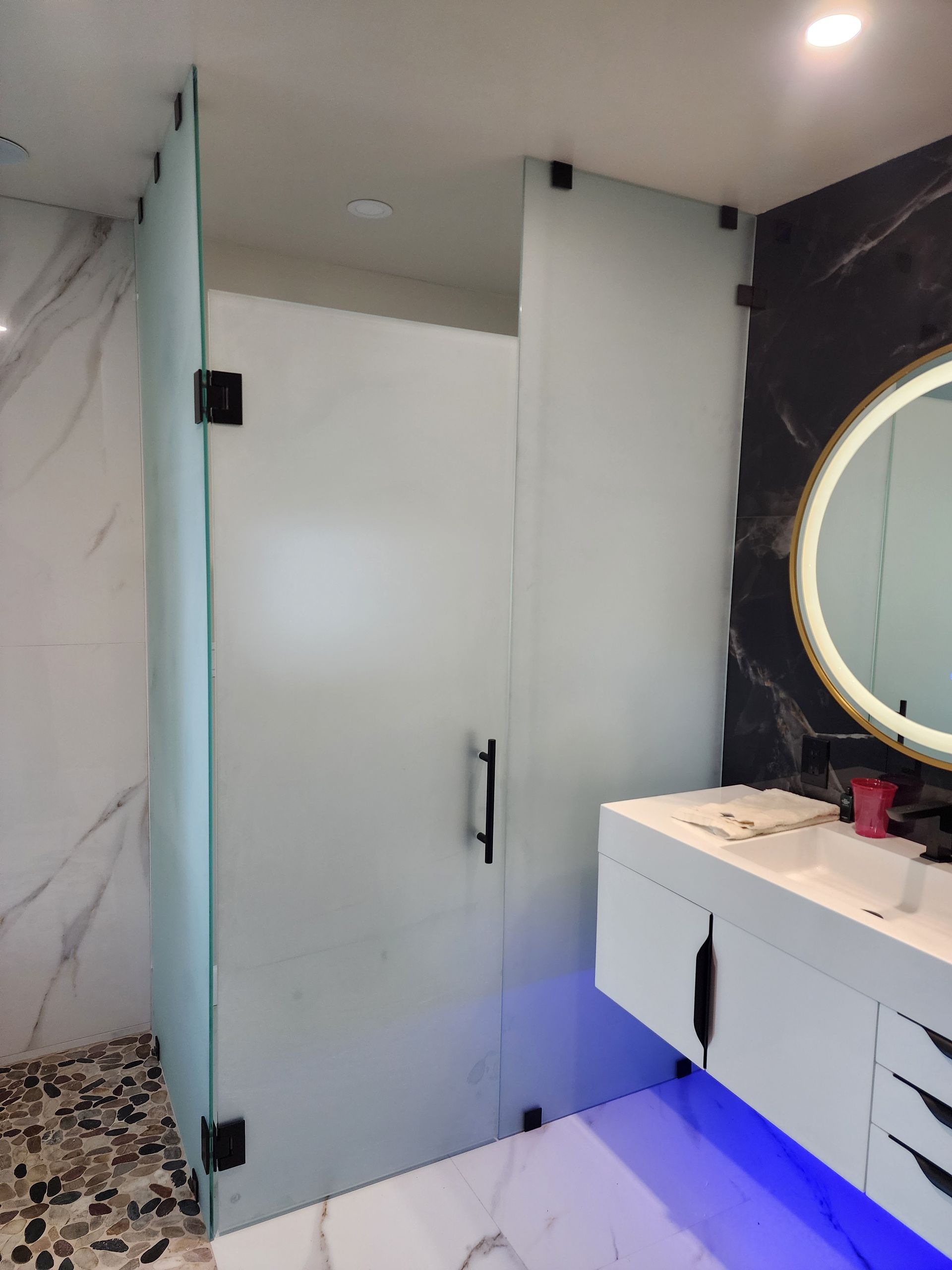 A frosted glass shower enclosure with matte black hardware sits next to a white vanity with blue under-cabinet lighting.