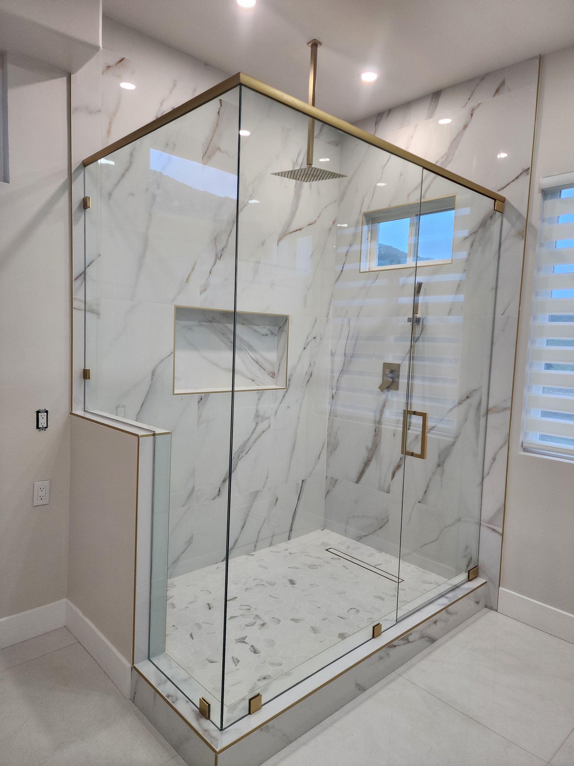 Modern glass shower stall with marble-patterned tile, brass hardware, and a rainfall showerhead in a bright bathroom.