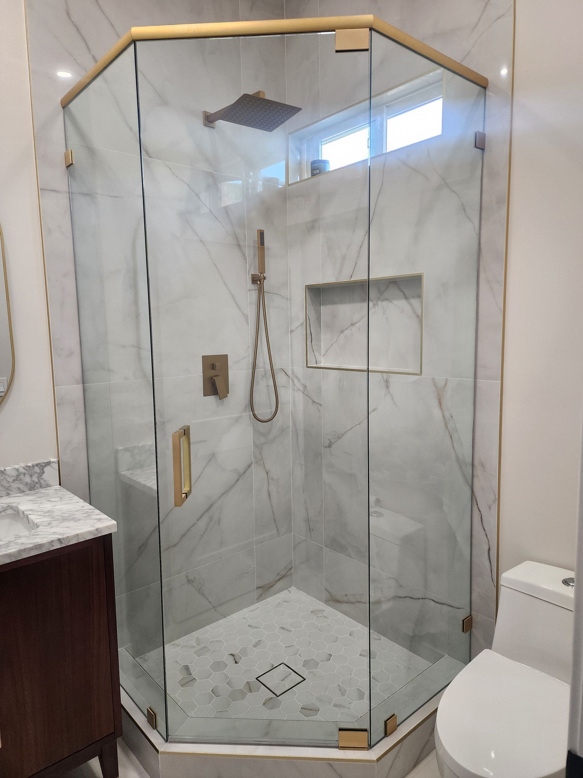 A corner shower with a glass enclosure, brass hardware, marble-patterned tile walls, and a rainfall showerhead.