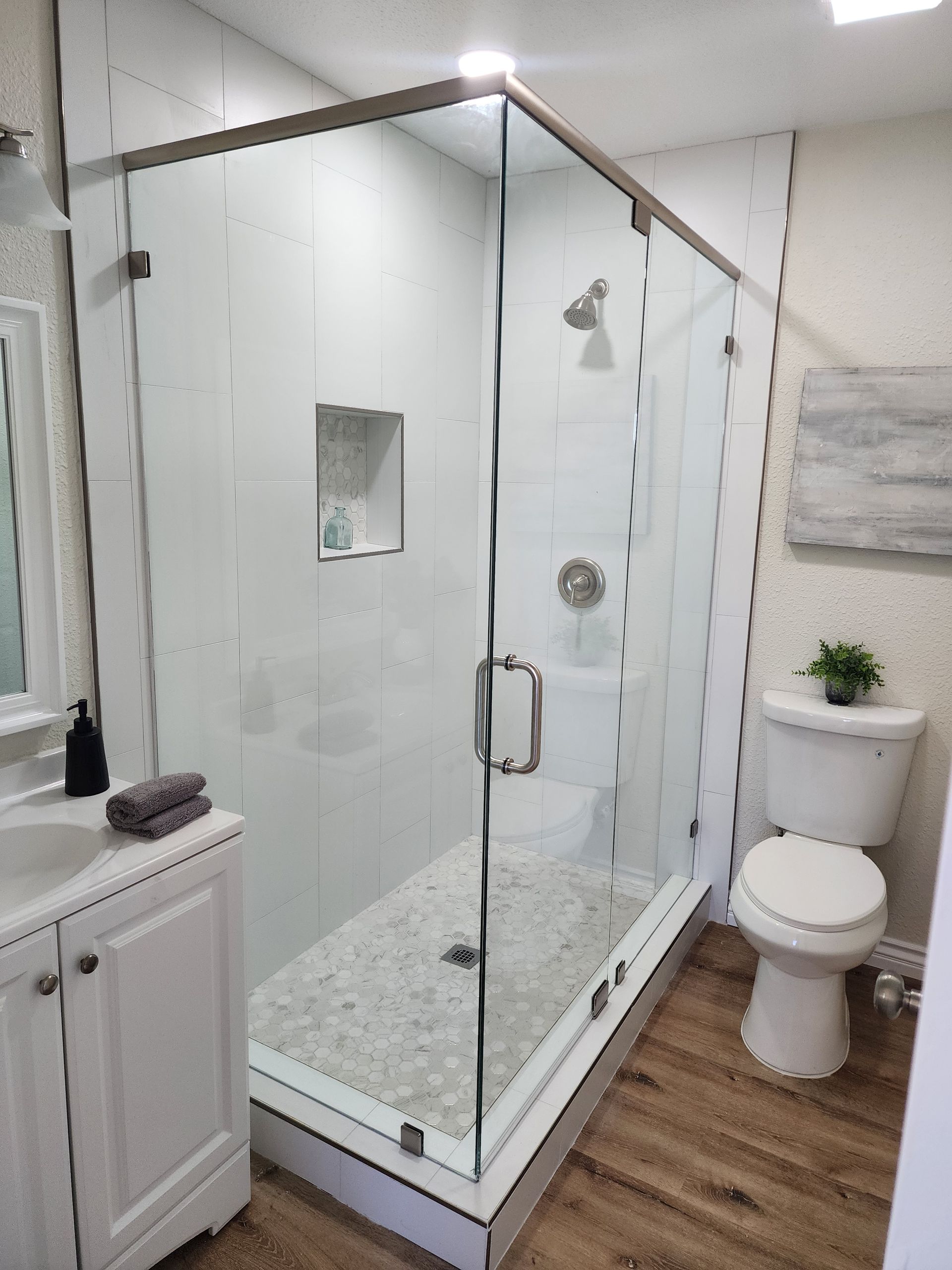 A modern bathroom featuring a corner glass-enclosed shower with pebble tile flooring, a white vanity, and a white toilet.